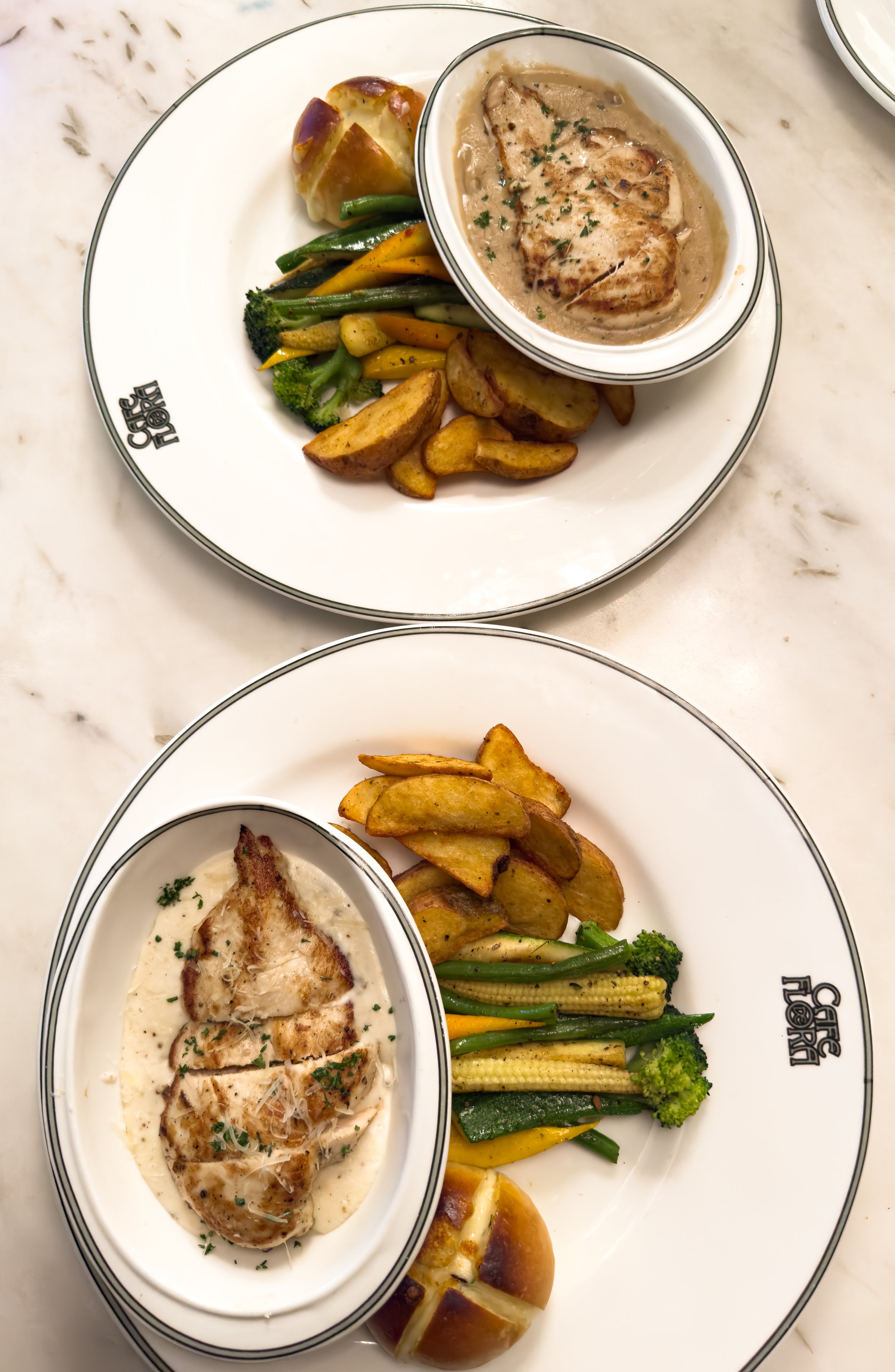 Two plates with grilled chicken, roasted potatoes, mixed vegetables, and bread with a creamy sauce, served on white plates with black rims.