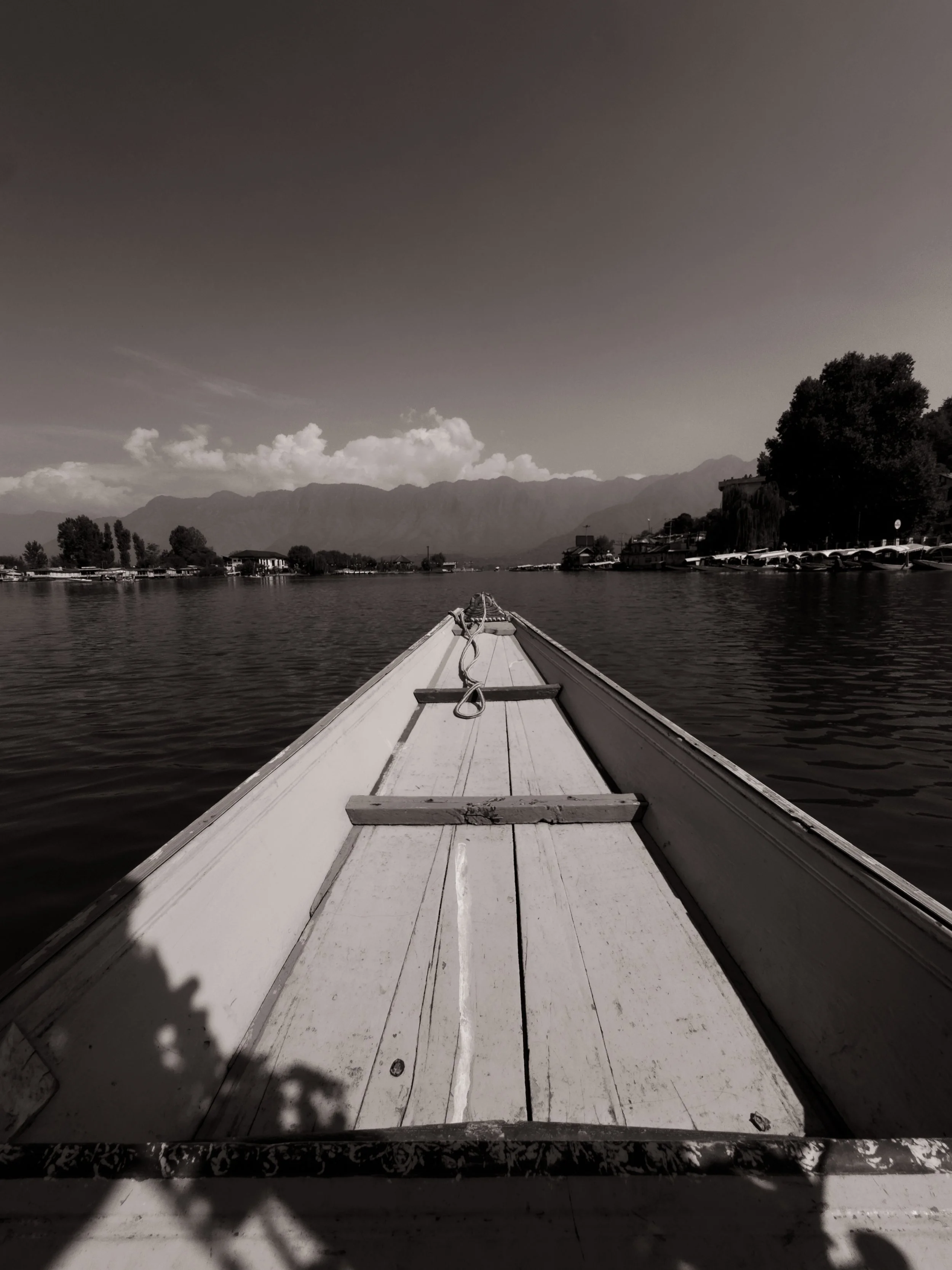 View from the front of a boat on a calm lake with mountains in the background, some trees on the shoreline, and parked boats or cars on the right side.