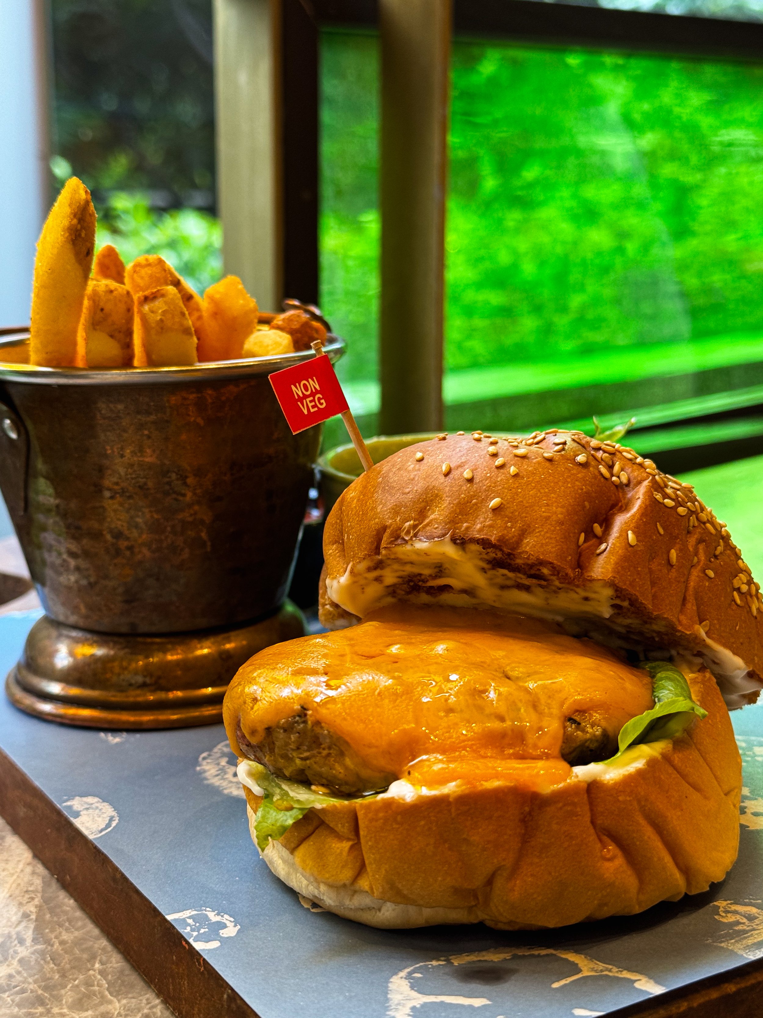 Cheeseburger with a sesame seed bun, lettuce, melted cheese, and a hamburger patty on a paper-lined tray, accompanied by a side of non-vegetarian fried potato wedges in a metal container.