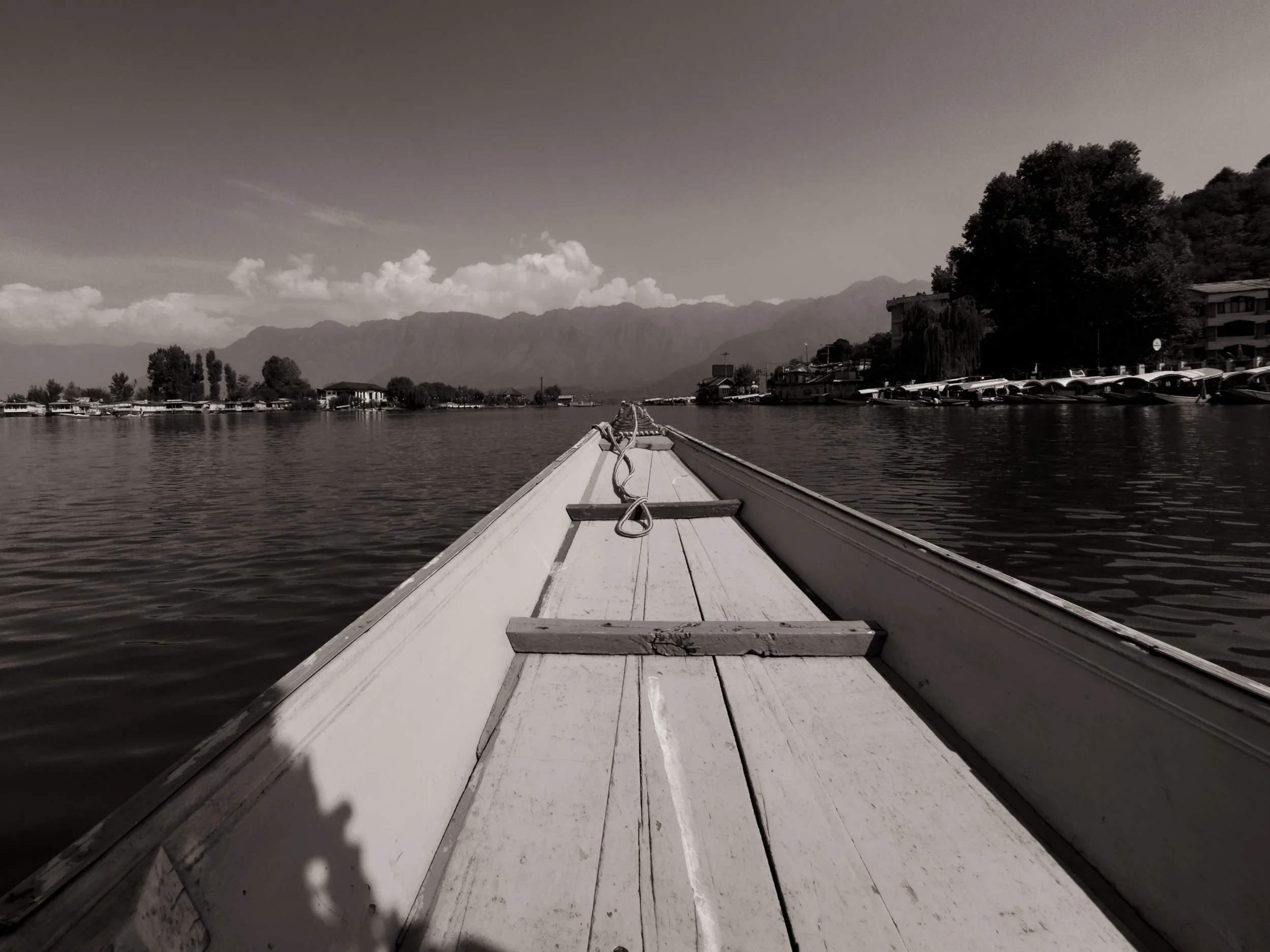 Black and white photo from the front of a boat on a lake, showing the boat's bow with ropes and wooden planks, calm water, distant islands, mountains, and partly cloudy sky.
