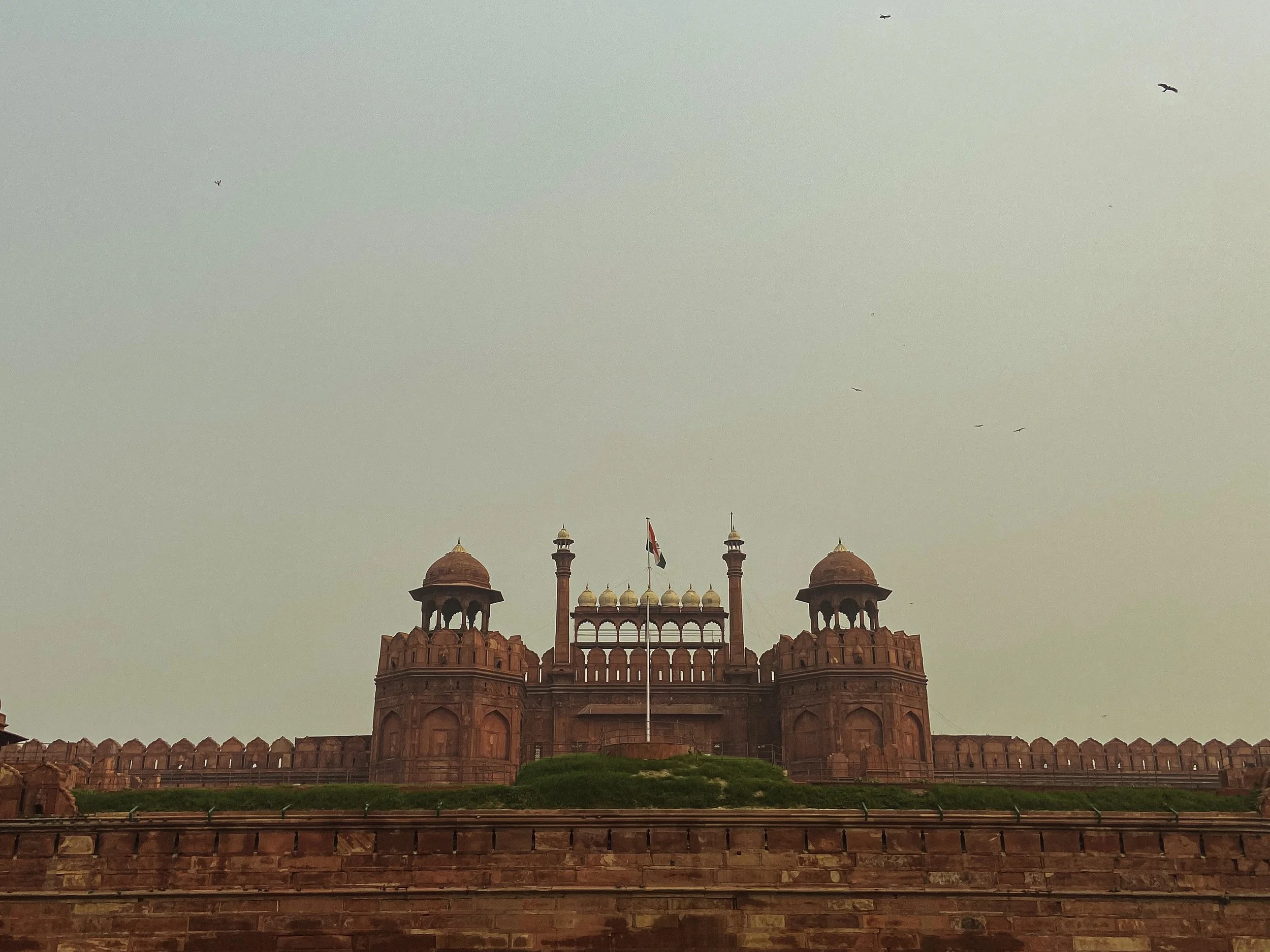 Red sandstone building with domes and minarets, and a flag in front, against a pale sky with some birds flying.
