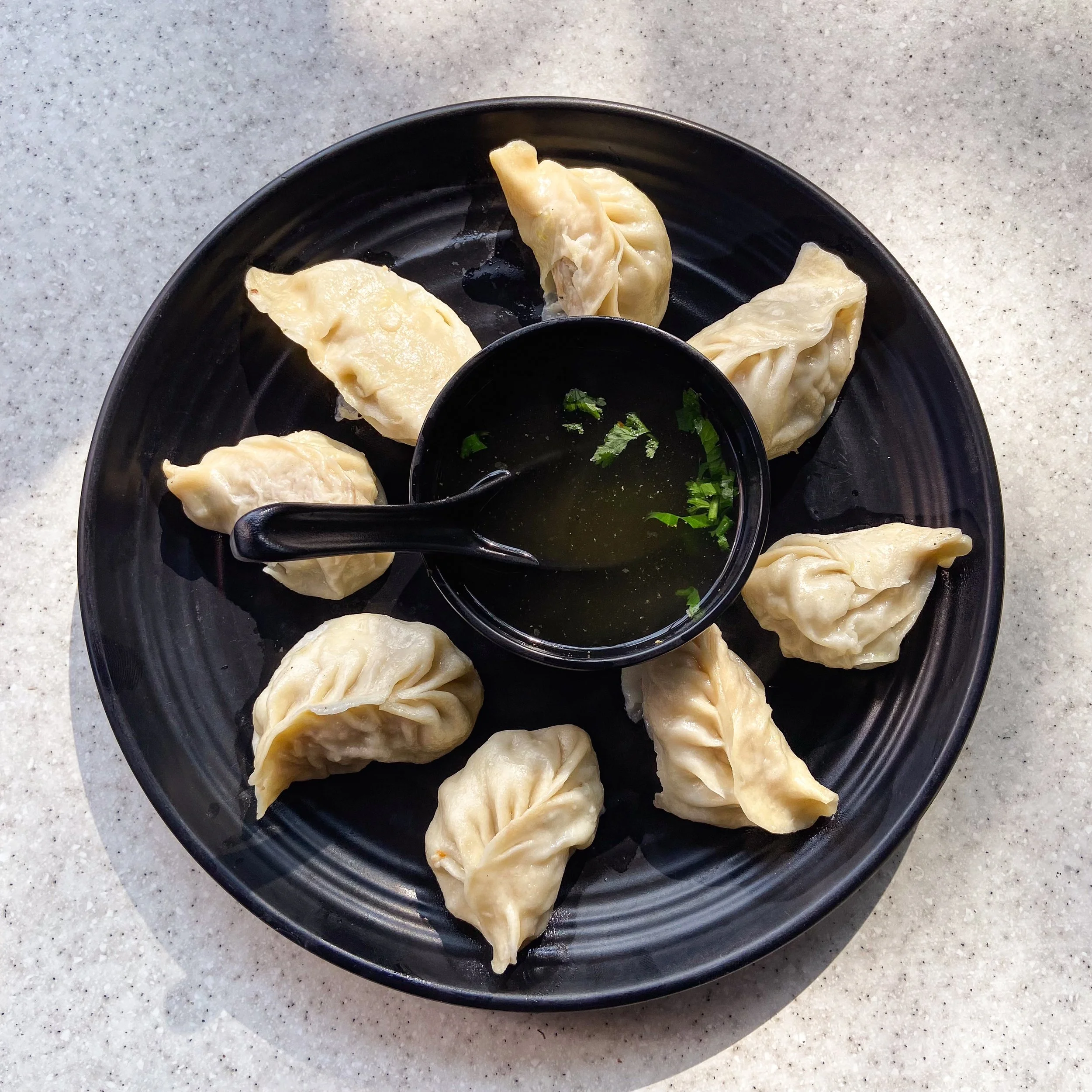Plate of eight steamed dumplings with a small bowl of soy sauce and chopped green herbs in the center on a white speckled surface.