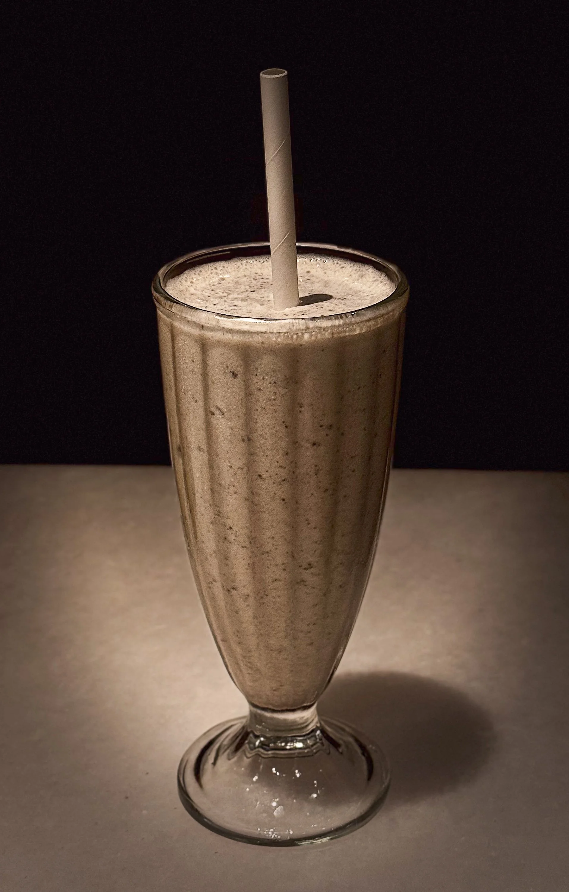 A tall glass of a frothy brown milkshake with a paper straw against a dark background.