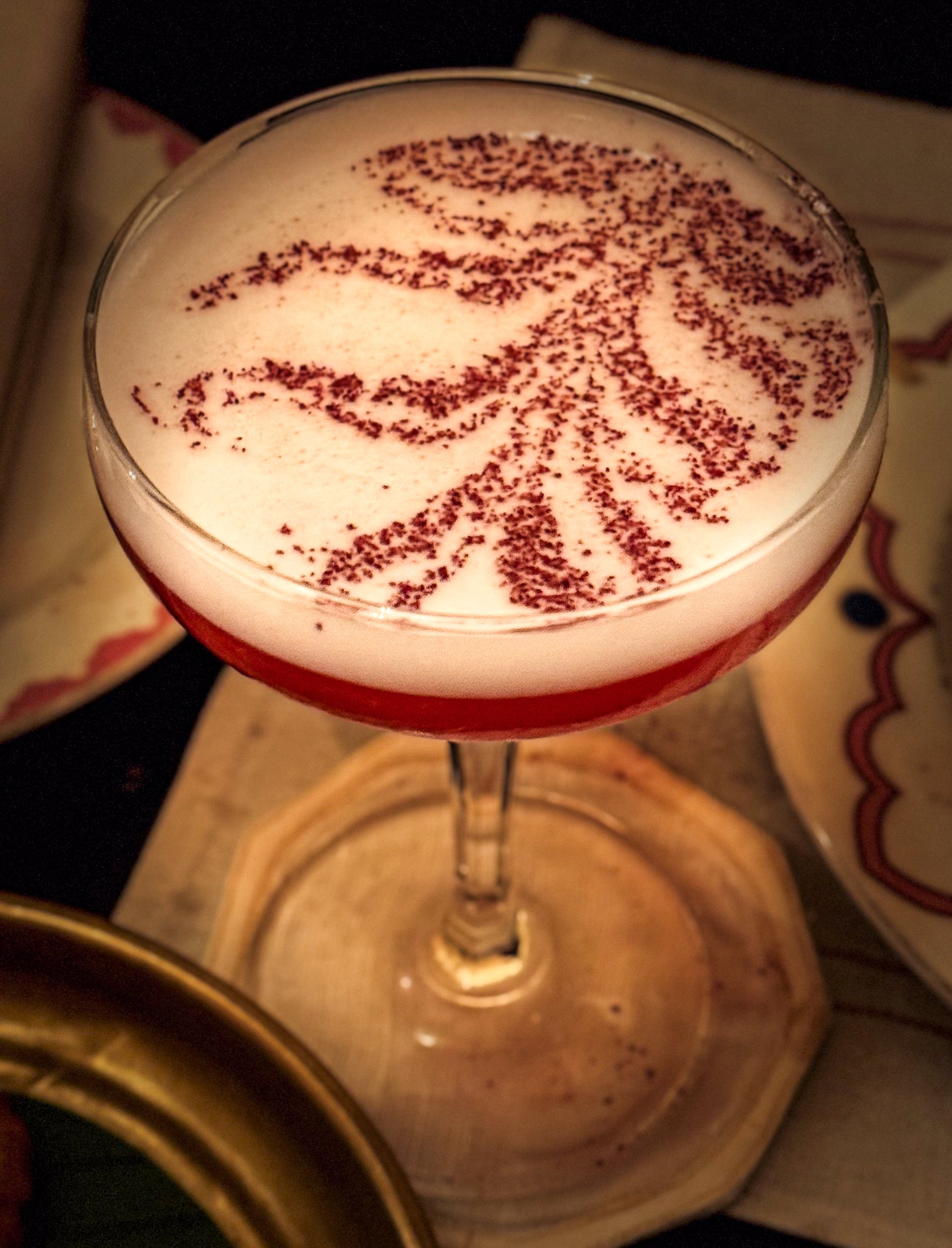 A cocktail in a coupe glass with a red layer at the bottom and white foam on top, decorated with red sprinkles in a floral pattern.