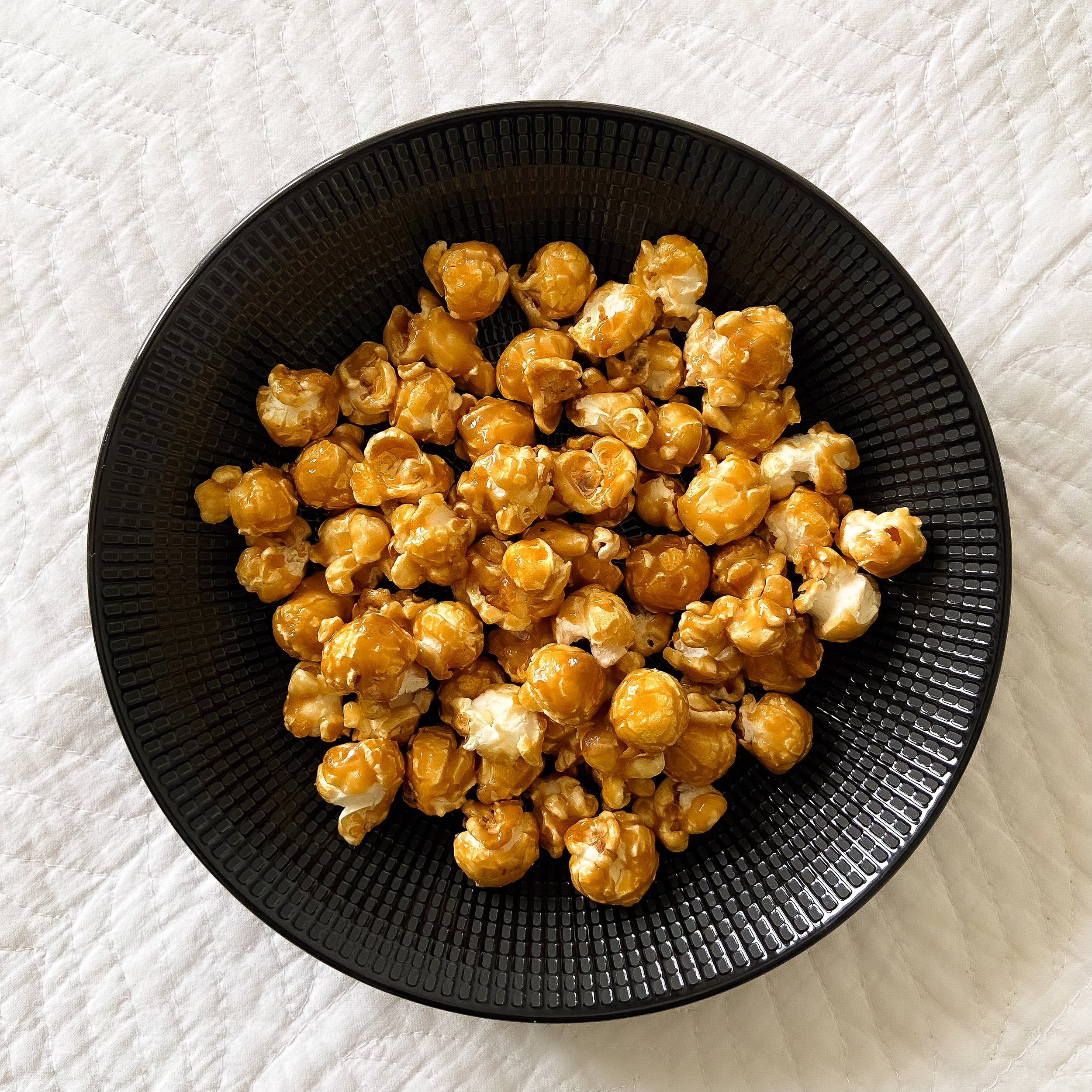 A black bowl filled with caramel popcorn placed on a white quilted fabric background.