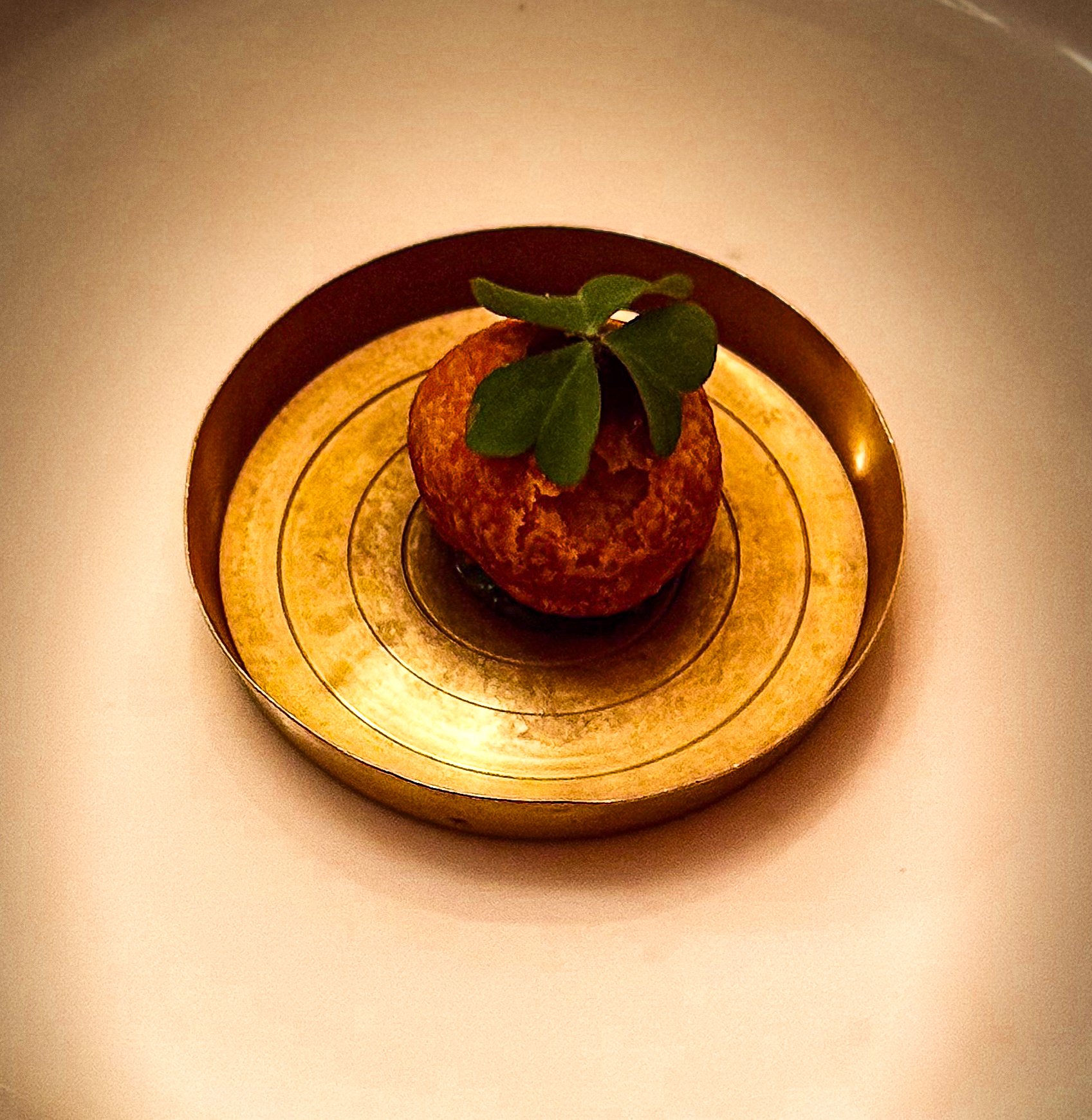 A small round pastry topped with green leaves on a gold-colored, reflective tray.