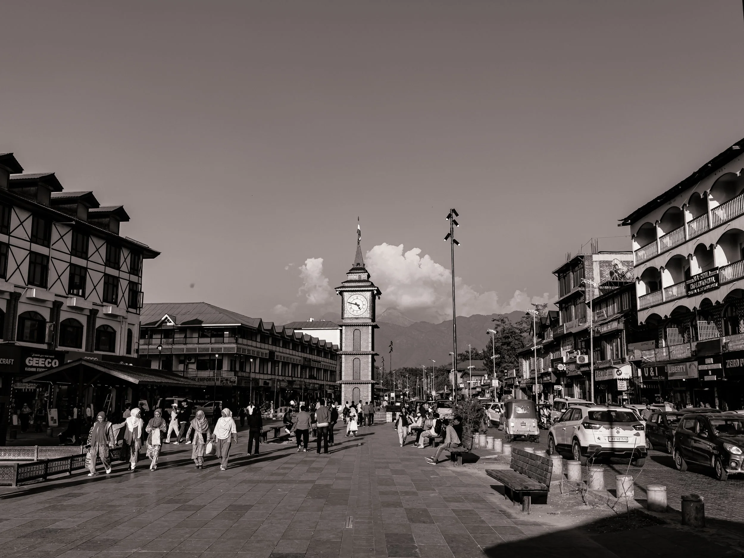 A street scene in a small town with a clock tower in the center, surrounded by buildings with shops and cafes. People are walking, sitting on benches, and cars are parked along the street. Mountain range is visible in the background.