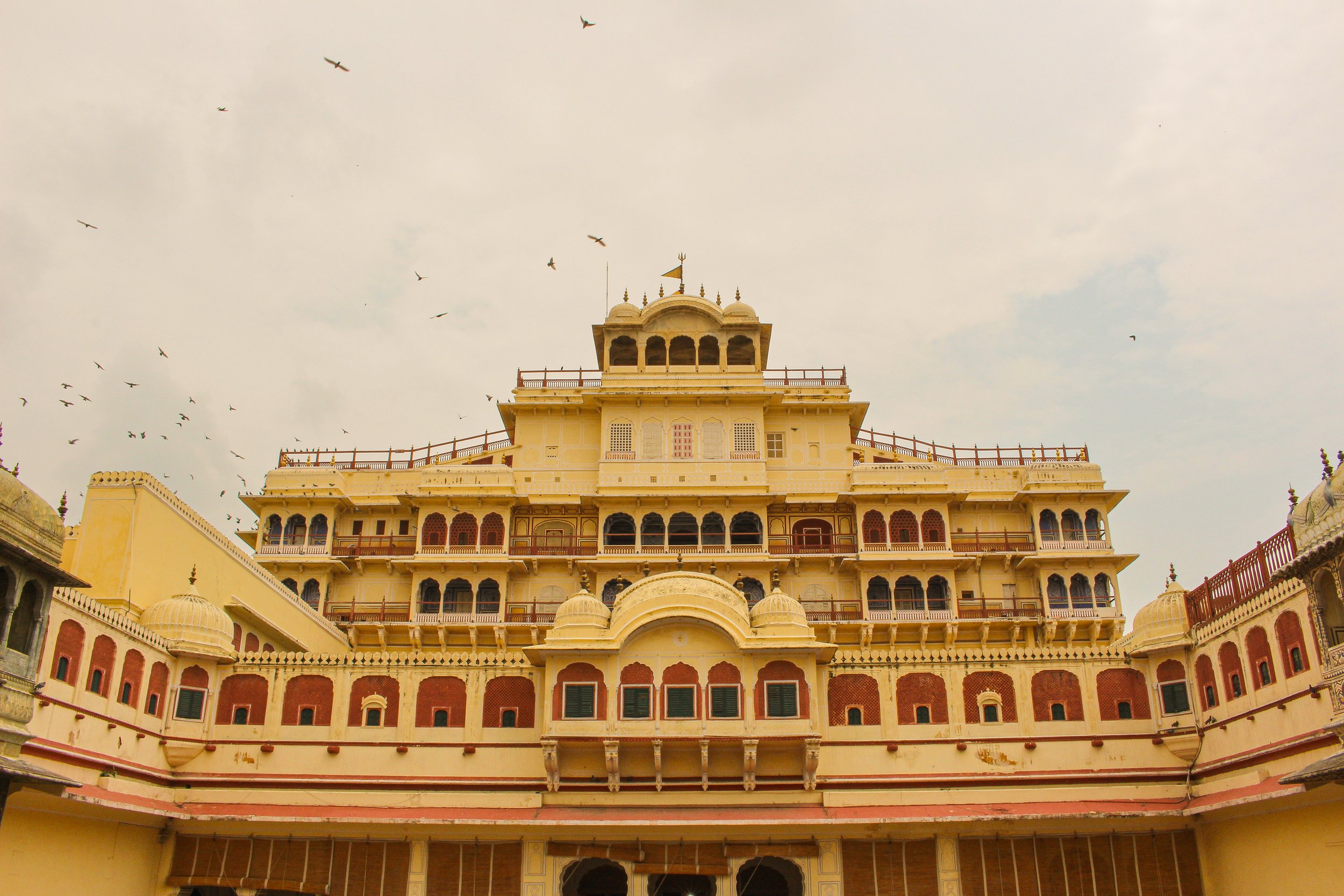 Front view of a traditional Indian palace with multiple levels, domes, and balconies, under a partly cloudy sky with birds flying overhead.
