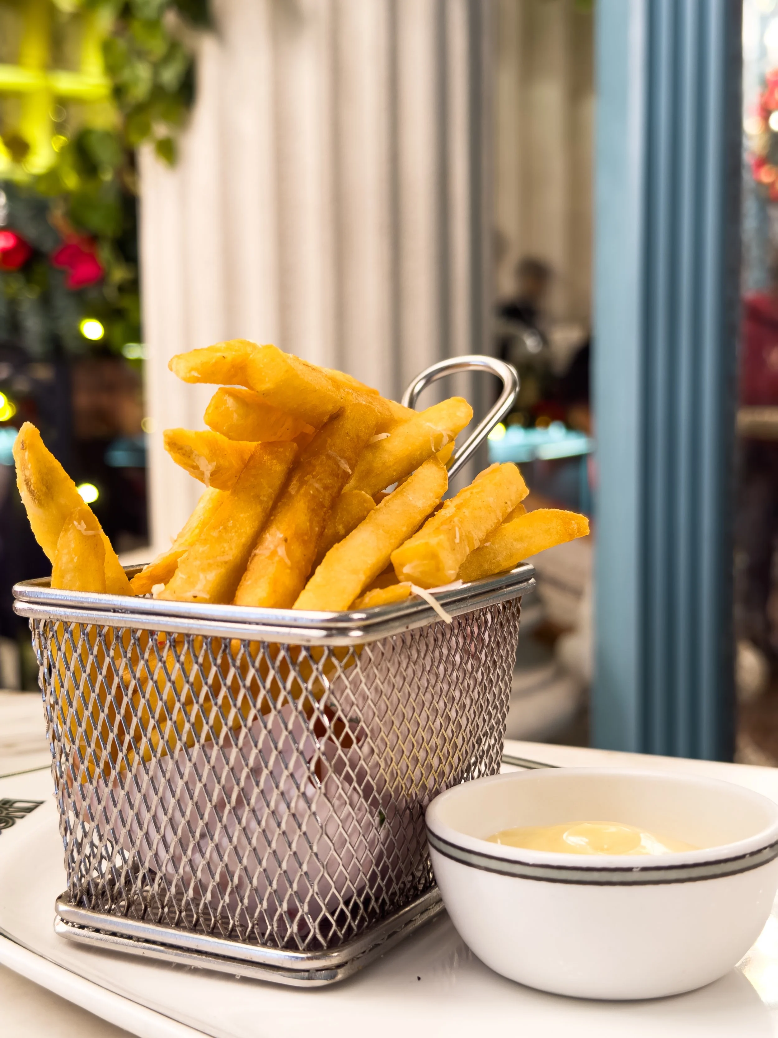 Basket of French fries with a small bowl of dipping sauce on a white plate in a restaurant setting.