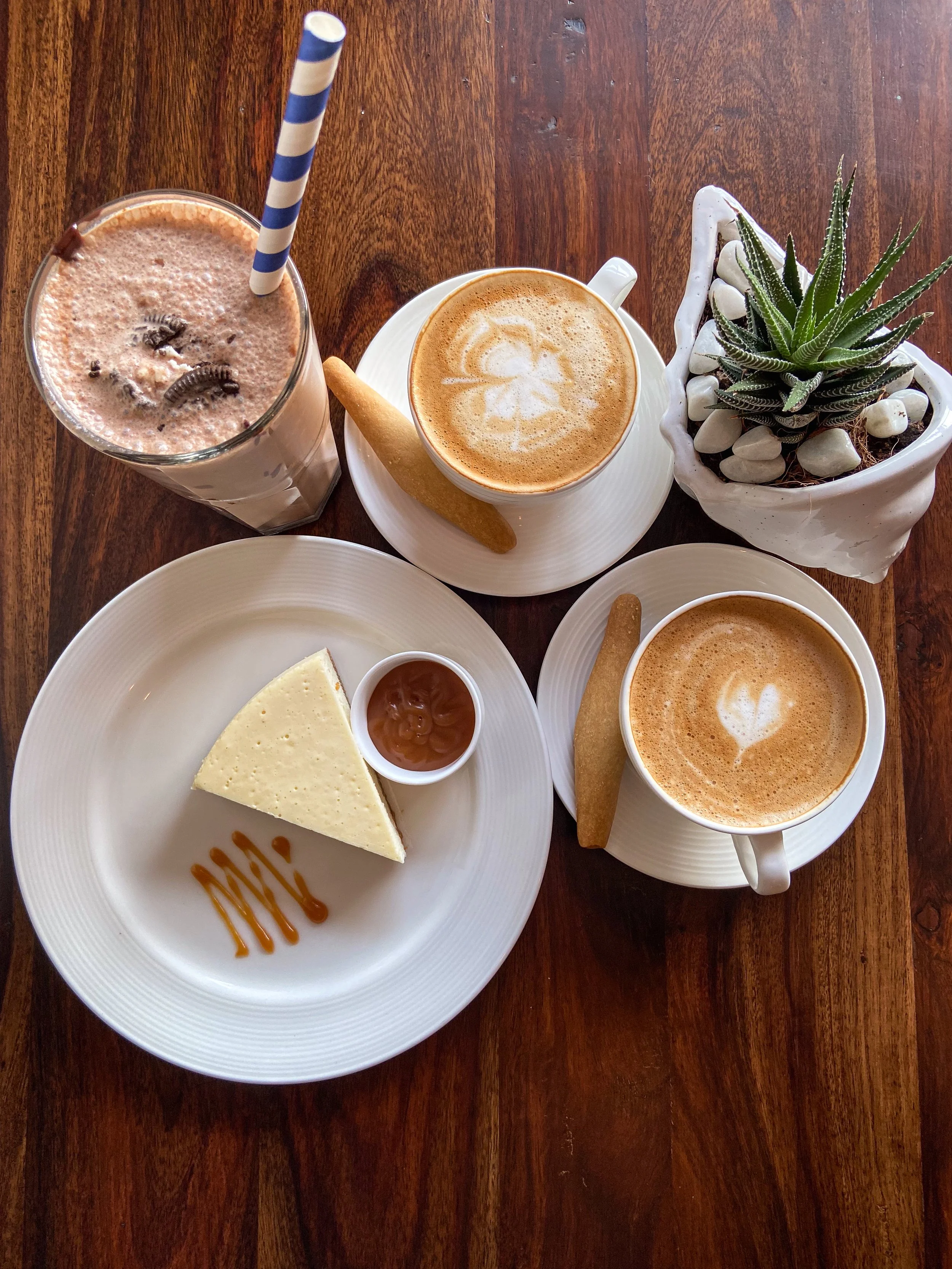 A dessert and coffee setup on a wooden table: a slice of cheesecake with caramel and a small cup of caramel sauce, two cups of coffee with latte art, a tall glass of chocolate milkshake with a striped straw, and a potted succulent plant in a white po
