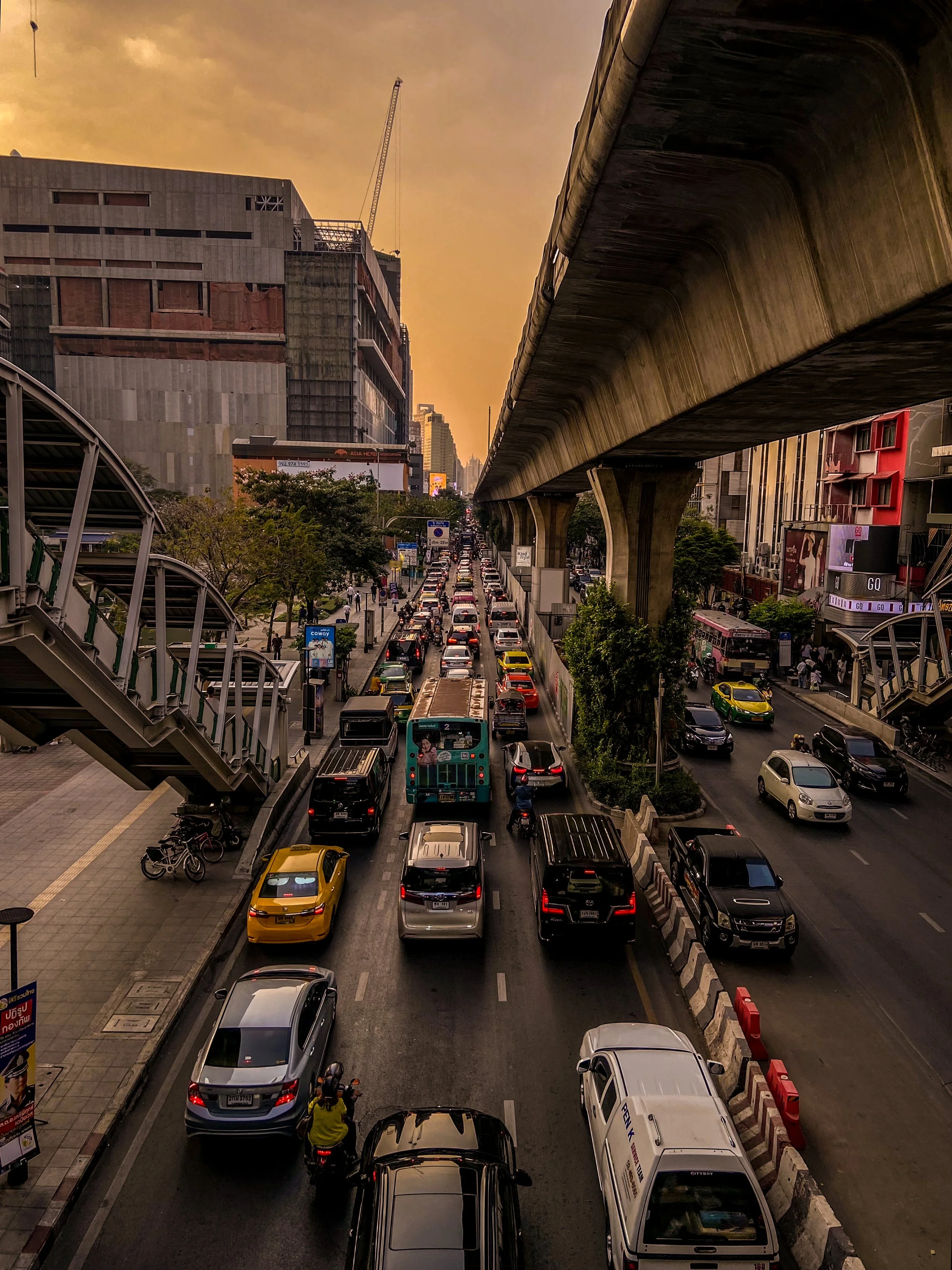 Traffic congestion on a city street with various cars and buses, elevated train tracks overhead, tall buildings on each side, and a golden sunset sky.