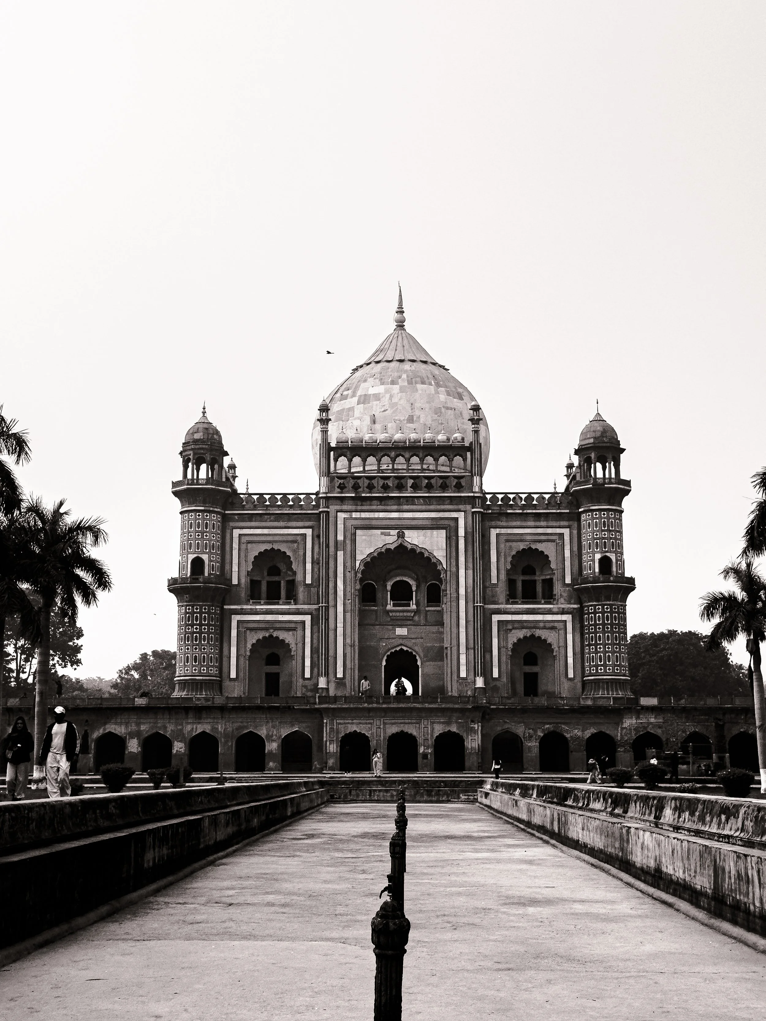 Black and white photo of the Taj Mahal with a pathway leading to the entrance, surrounded by trees and visitors.