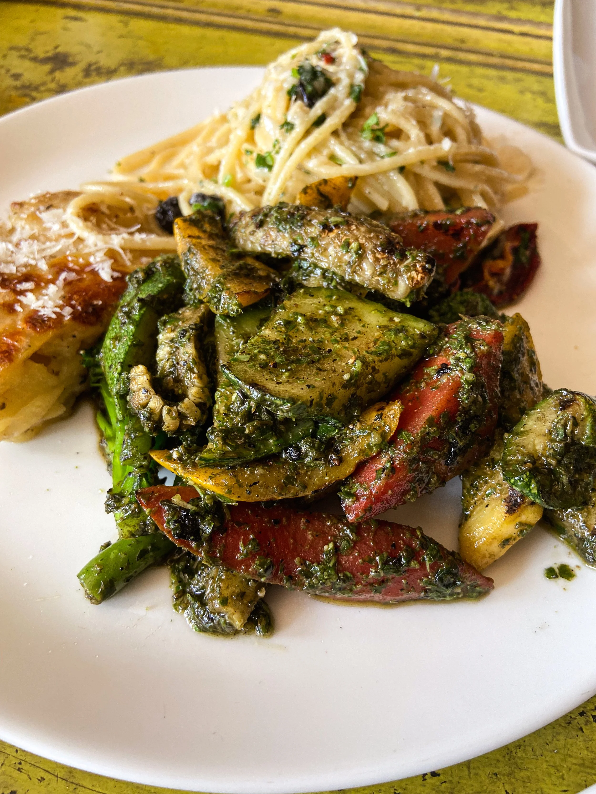 Plate of grilled vegetables with herbs, pasta with spinach, and a slice of pizza, on a wooden table.