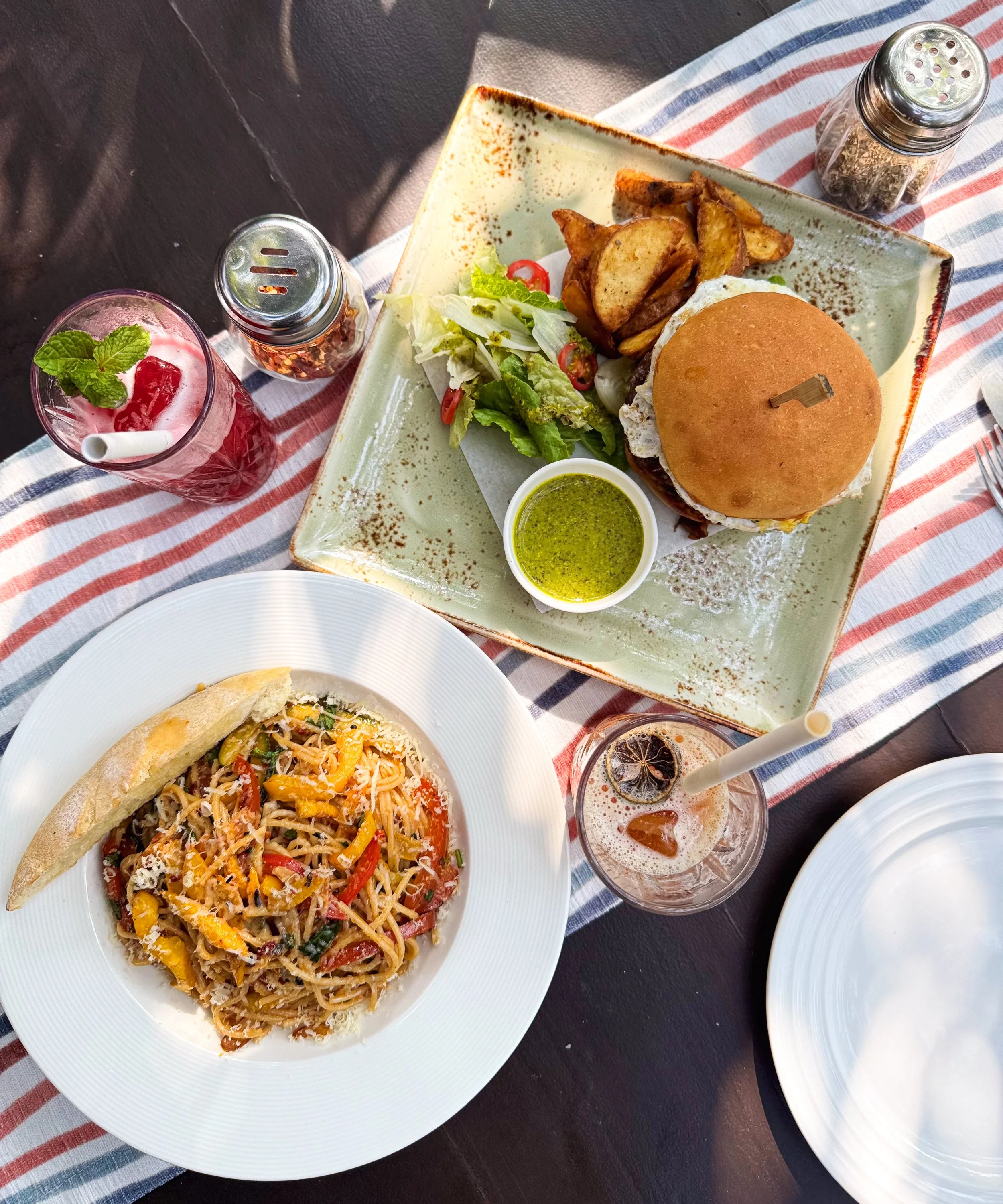 A spread of food and drinks on a table, featuring a burger with fries, a plate of pasta, a green salad, and beverages including iced tea and a pink drink.