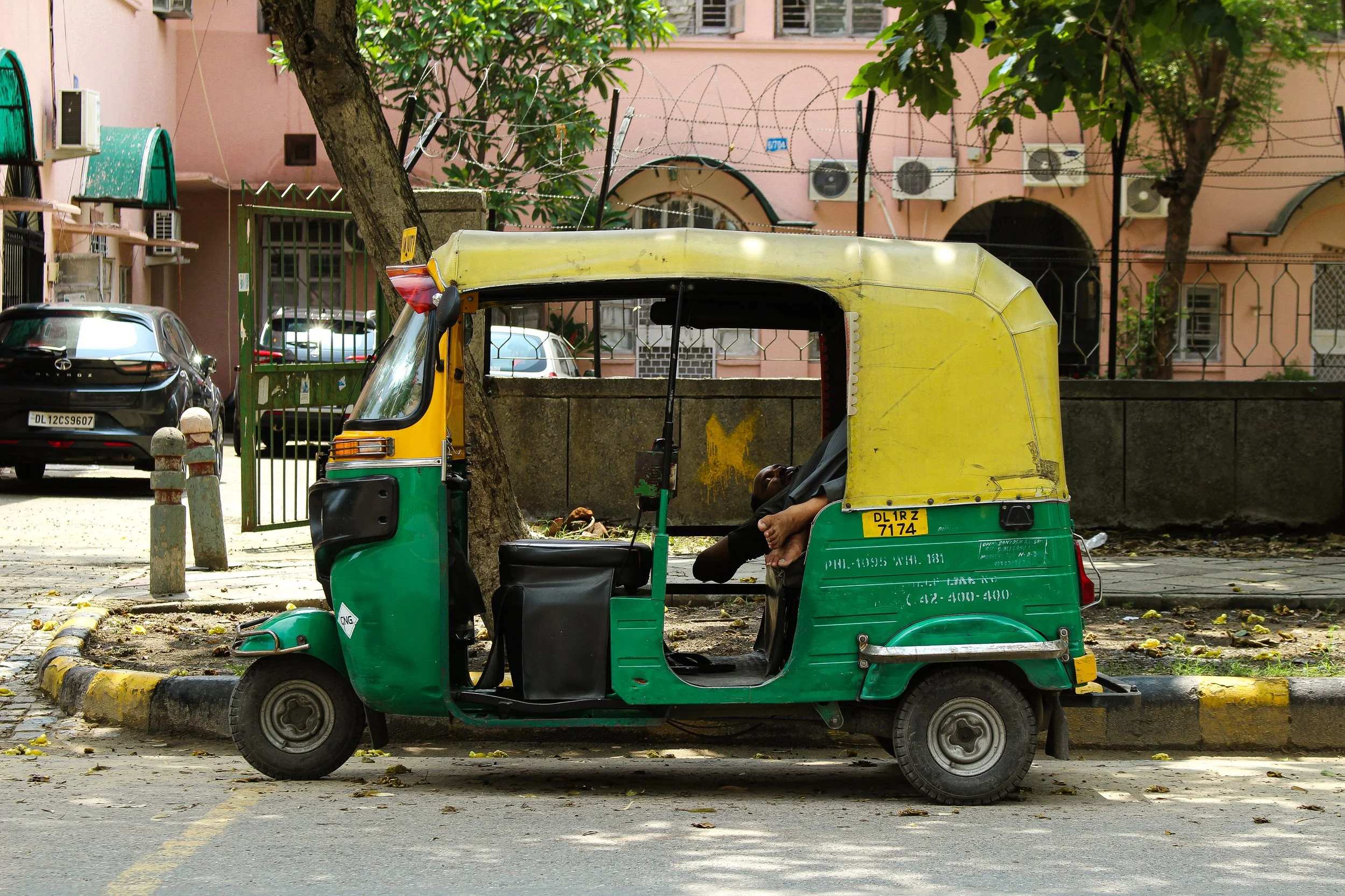 A parked green and yellow auto rickshaw on a city street with a man sleeping inside, leaning back with head tilted to the side. There are trees, cars, and pink buildings in the background.