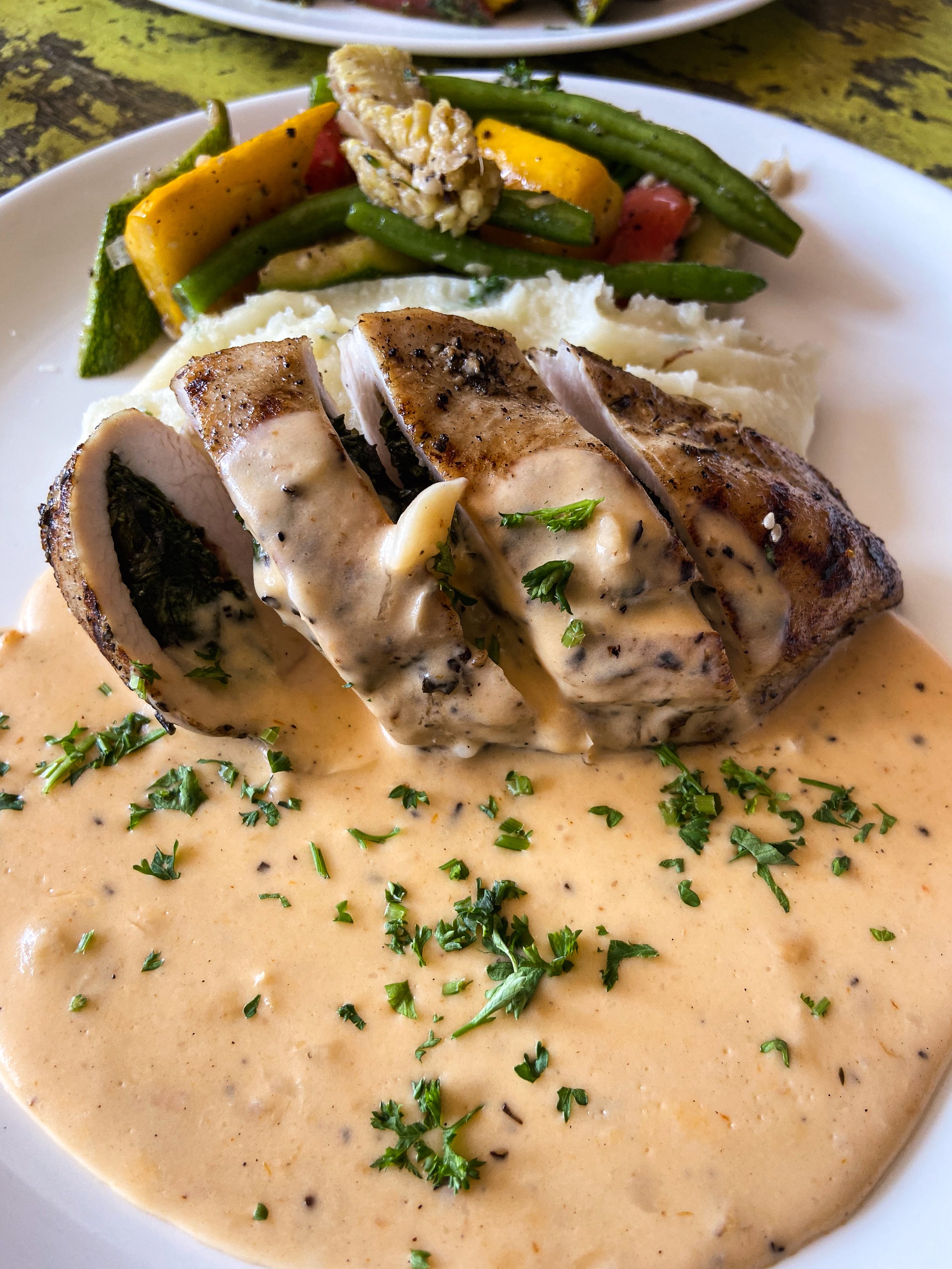 A plate of food with sliced chicken topped with creamy sauce, mashed potatoes, and mixed vegetables including green beans, yellow squash, zucchini, and cherry tomatoes.