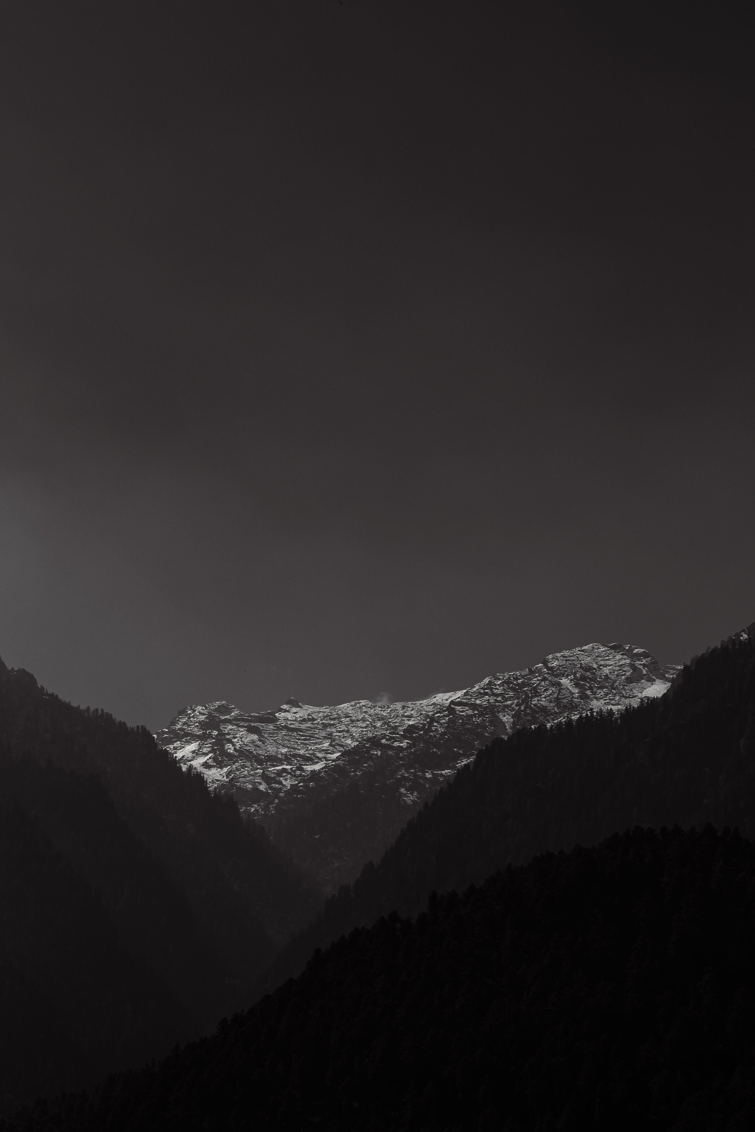 Black and white photograph of snow-capped mountains with dark silhouettes of forested hills in the foreground.