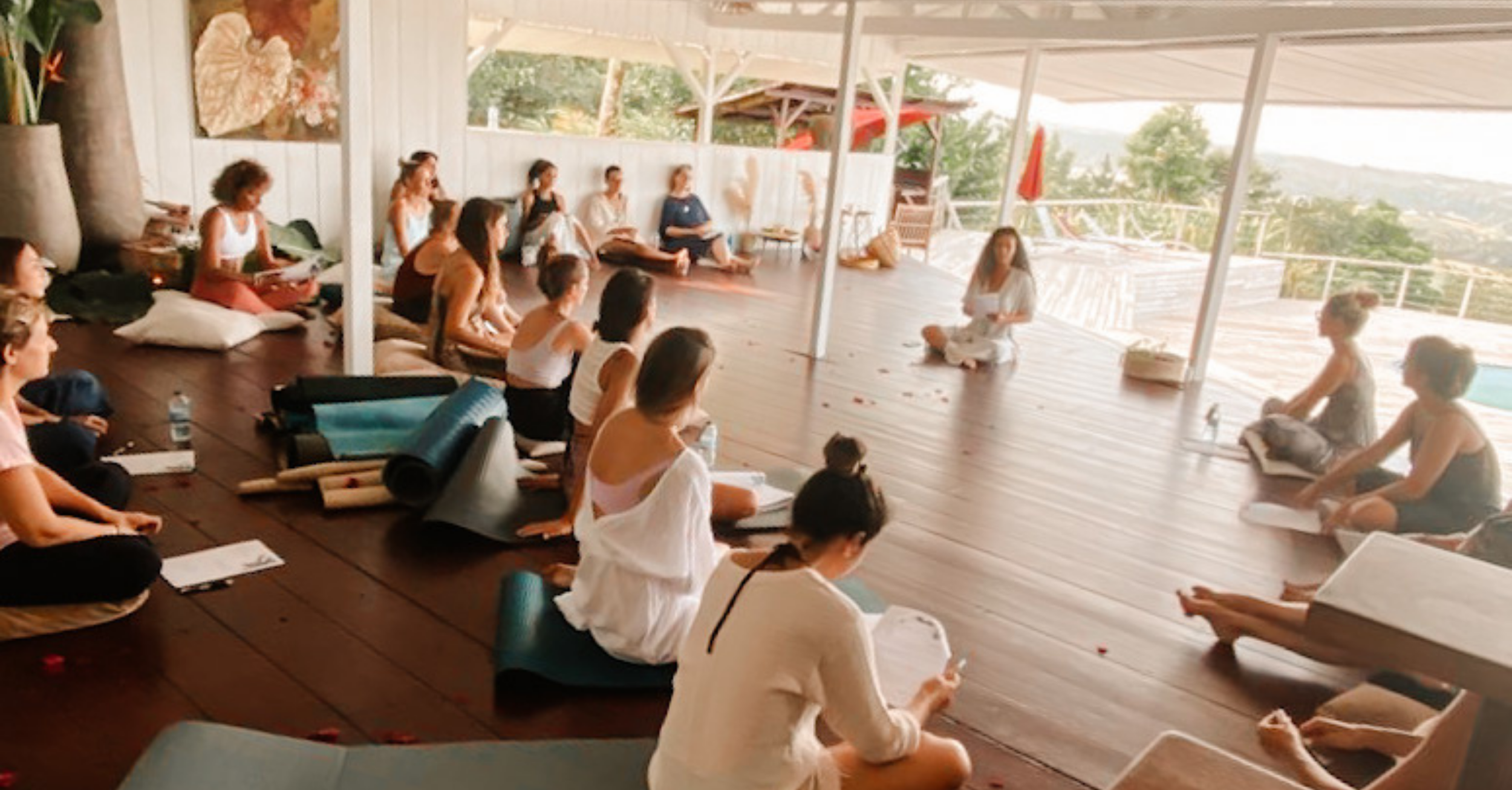 Groupe de personnes participant à un cours de méditation en position assise sur le sol en extérieur sous un toit en bois avec vue sur la nature.