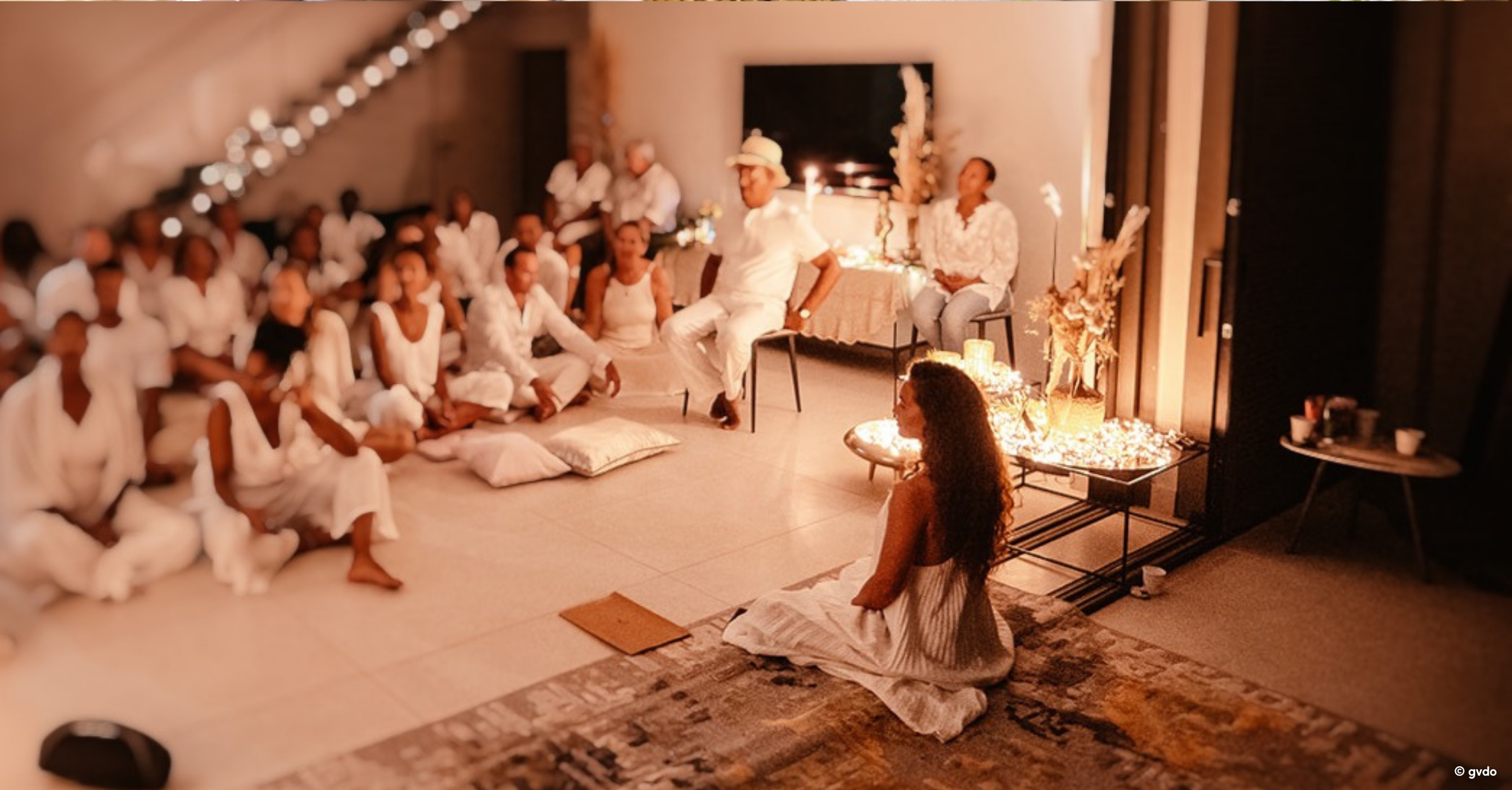 Une jeune femme assise à genoux sur un tapis, au centre d'un groupe de personnes qui semblent participer à une cérémonie ou un rassemblement. Le décor est chaleureux avec des bougies et des décorations florales, ambiance intime et spirituelle.