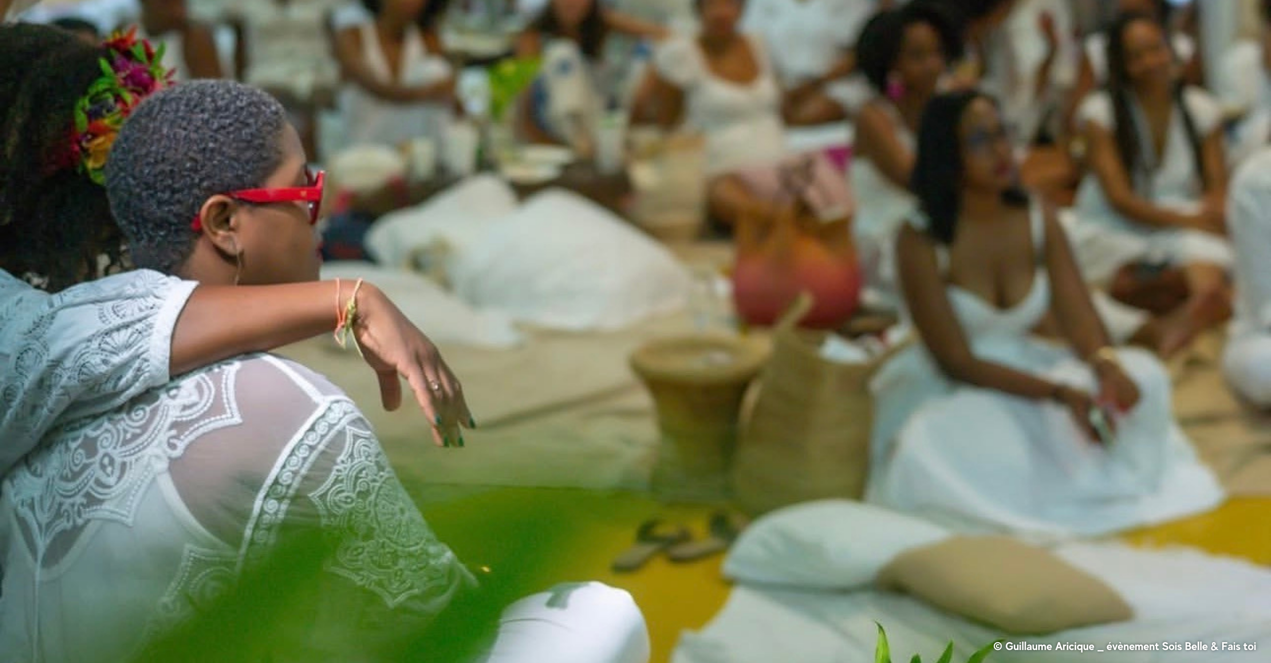 Groupe de femmes assises à terre lors d'un événement, portant des vêtements blancs et colorés, dans un environnement intérieur accueillant.