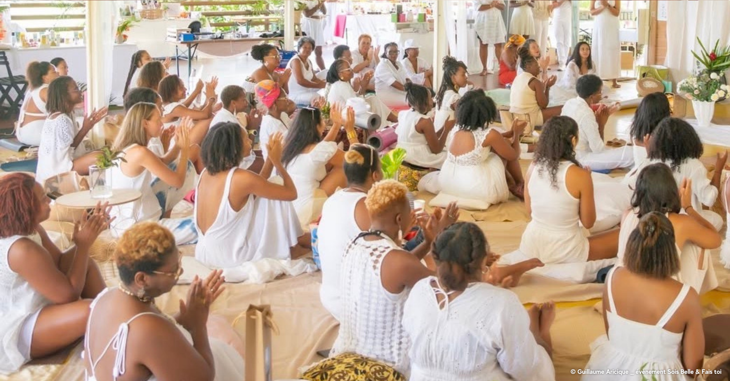 Groupe de femmes et filles assises par terre en méditation ou prière dans une salle lumineuse décorée de plantes et fleurs, portant principalement des vêtements blancs.