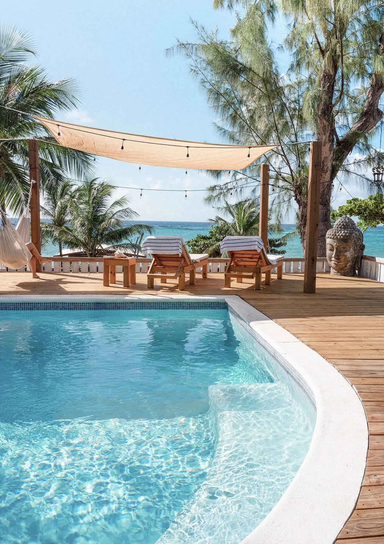 Poolside view at a tropical beach resort with lounge chairs, a canopy, palm trees, ocean in the background, and a stone Buddha head sculpture.