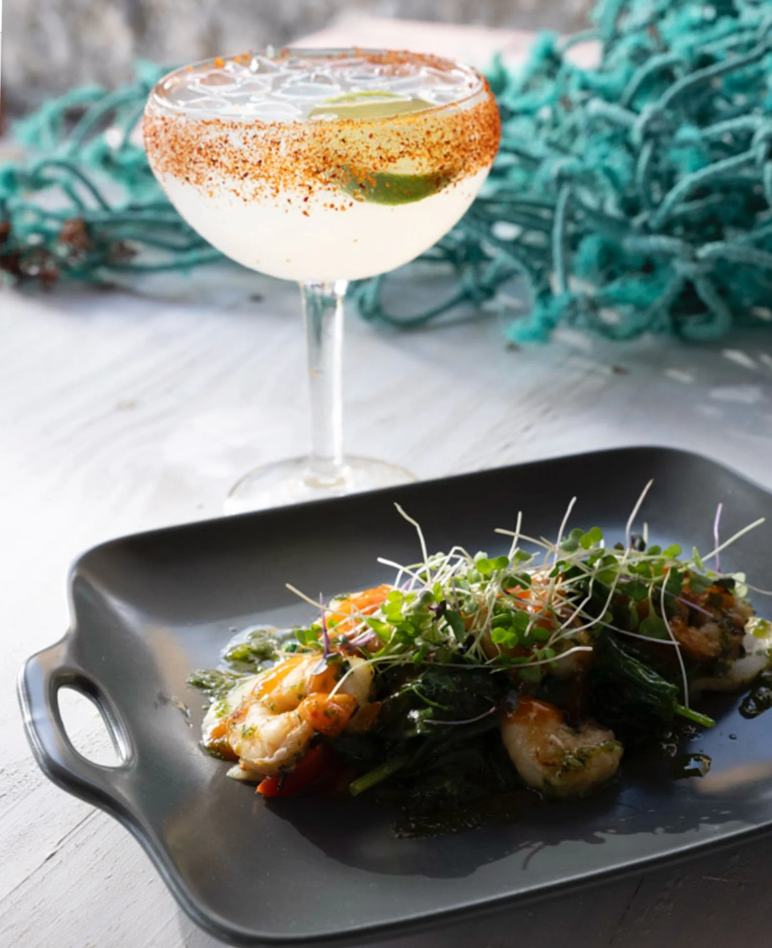 A silver serving tray with grilled shrimp, vegetables, and microgreens, served on a black plate. In the background, there is a martini glass with a white cocktail garnished with lime and chili powder.