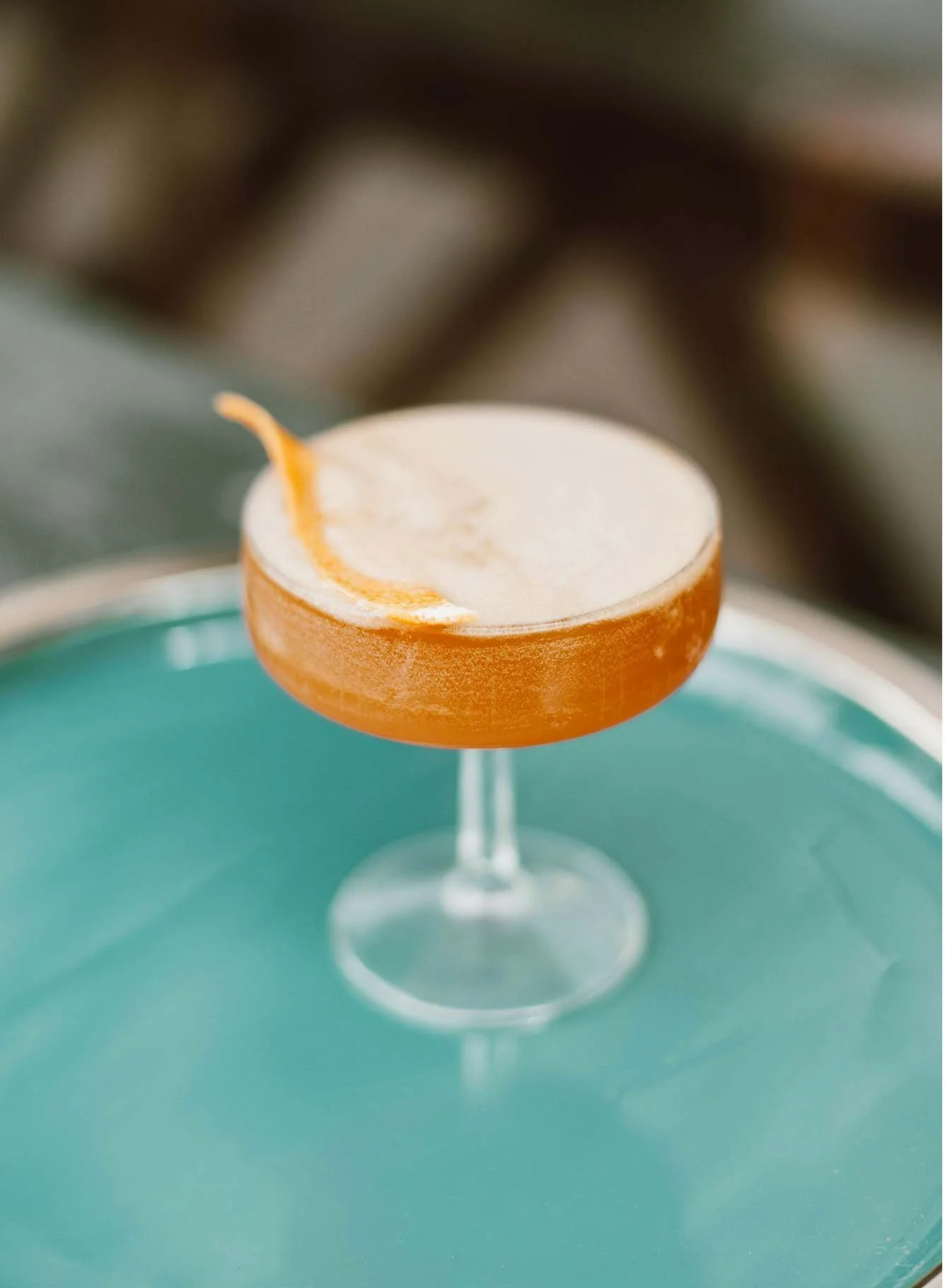 A cocktail glass with a frothy drink and a twist of lemon peel garnish, placed on a turquoise surface.
