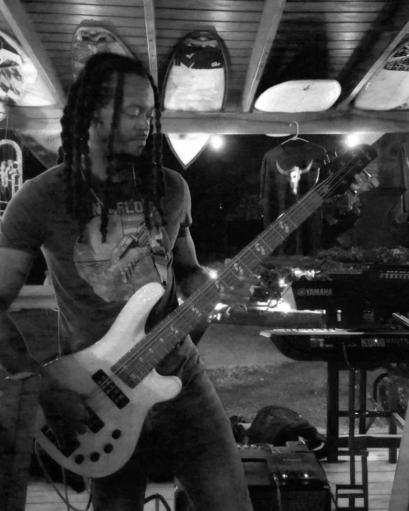 A person playing a bass guitar at night in a venue with surfboards on the ceiling and musical equipment around.