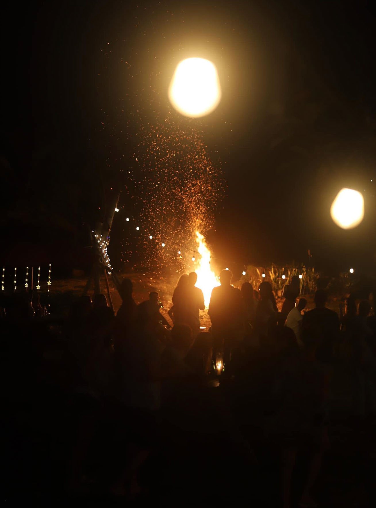 People gathered around a bonfire at night, releasing floating lanterns into the sky, with dark sky and stars in the background.