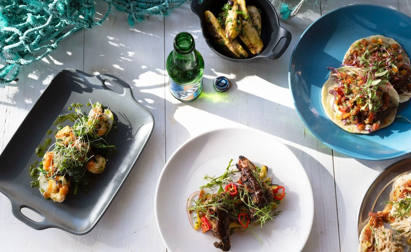 A variety of dishes on a white table, including tacos with lettuce, tomato, and sprouts in a blue bowl; grilled chicken drumsticks with chili peppers on a white plate; potato wedges in a black skillet; a black serving tray with skewered shrimp garnished with microgreens and green sauce; and a green glass bottle of beer.