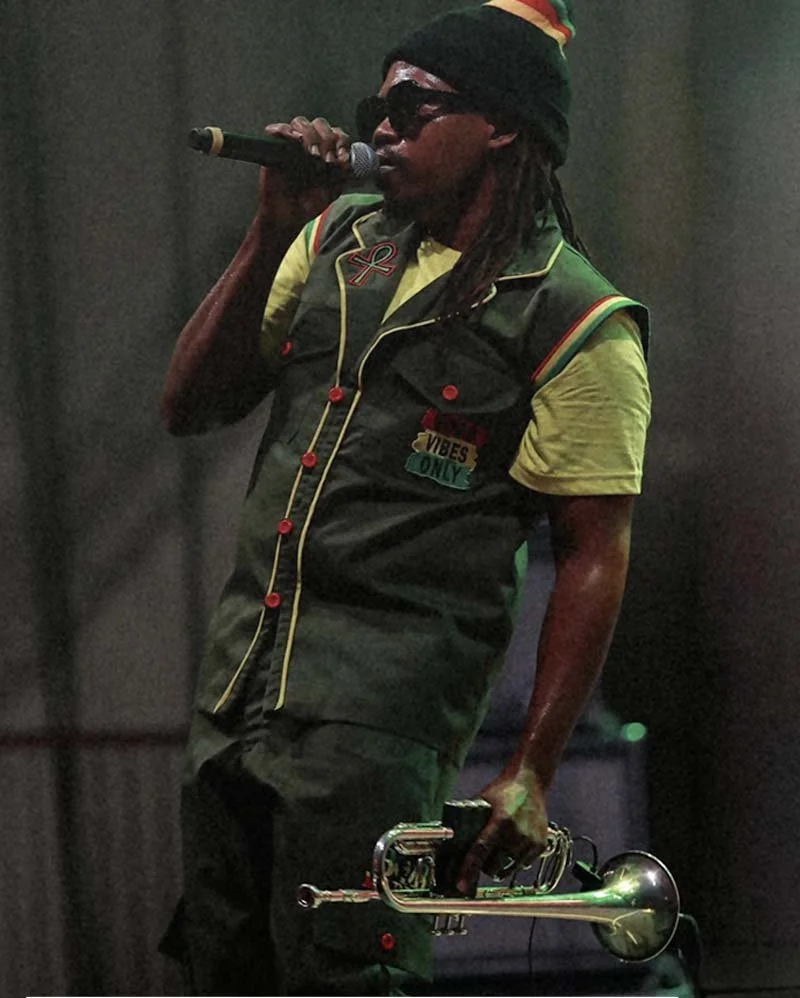 Person with dreadlocks wearing sunglasses, a colorful hat, and a sleeveless vest with patches, singing into a microphone and holding a trumpet.