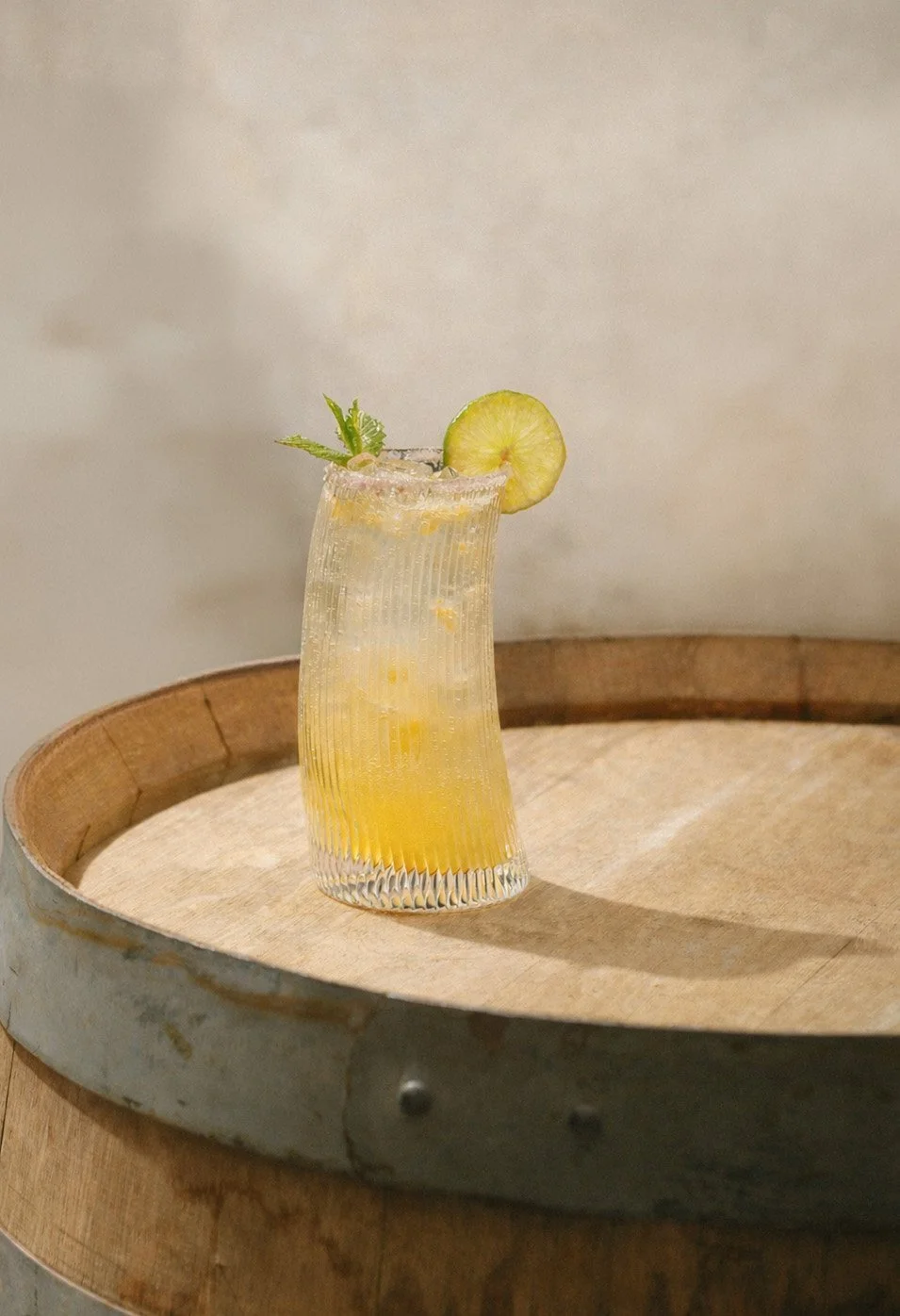 A glass of yellow beverage with a lemon slice and mint leaves on top, placed on a wooden tray.