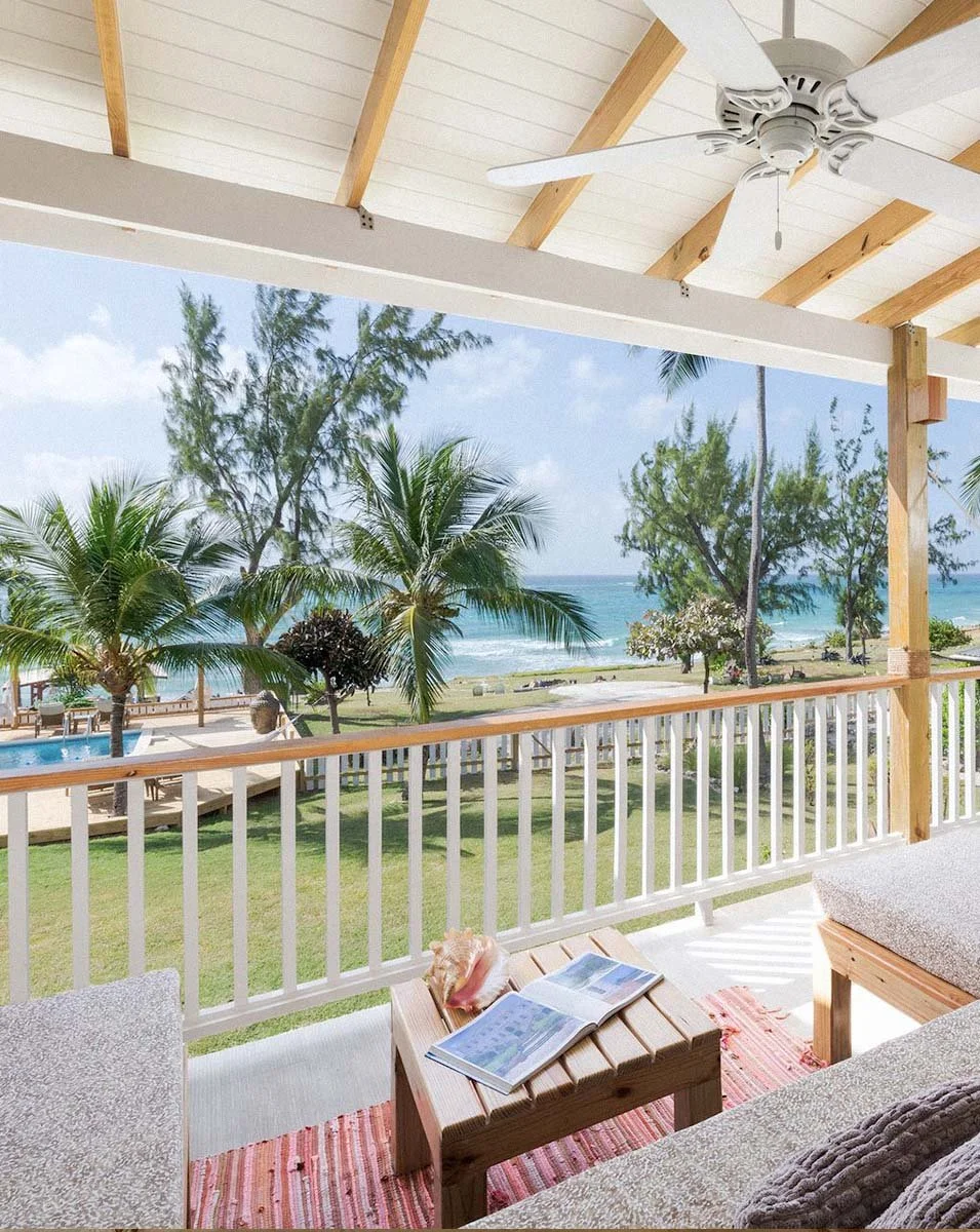 View from a covered porch with wooden ceiling and ceiling fan overlooking a tropical beach with palm trees, green grass, and ocean waves.
