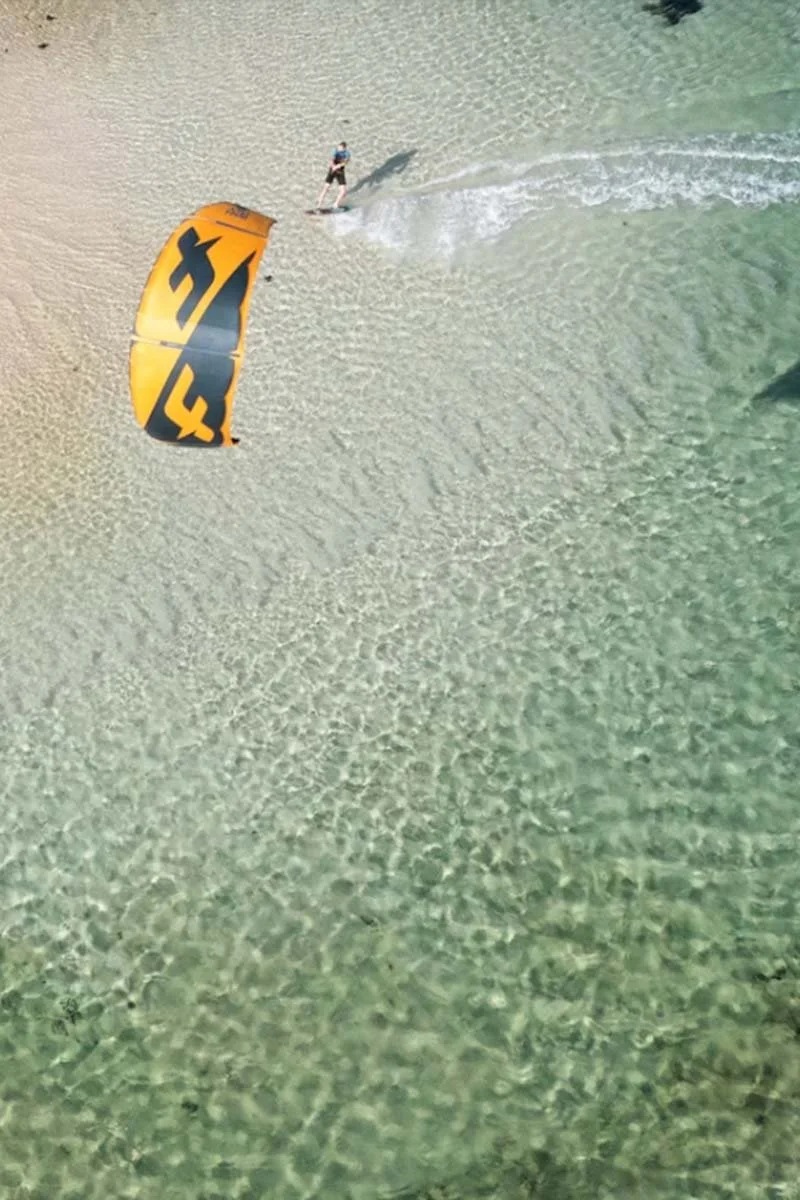 A person is kiteboarding on clear, shallow water near a sandy beach, holding onto a yellow and black kite that is partially inflated.