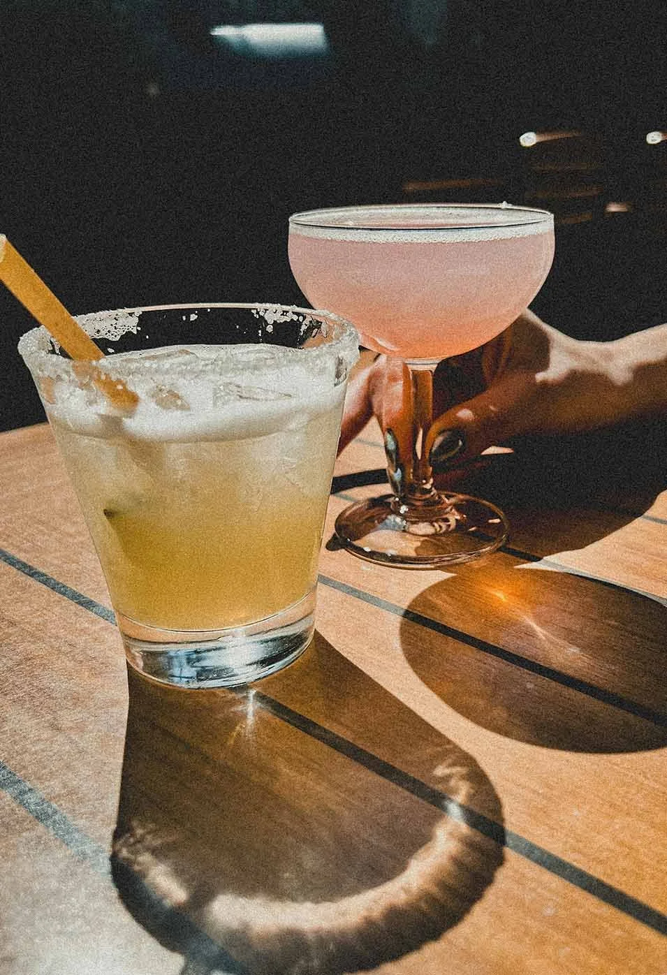 A yellow cocktail with ice and a straw in a clear glass, and a pink cocktail in a coupe glass, with a hand holding the pink drink, on a wooden table with shadows.