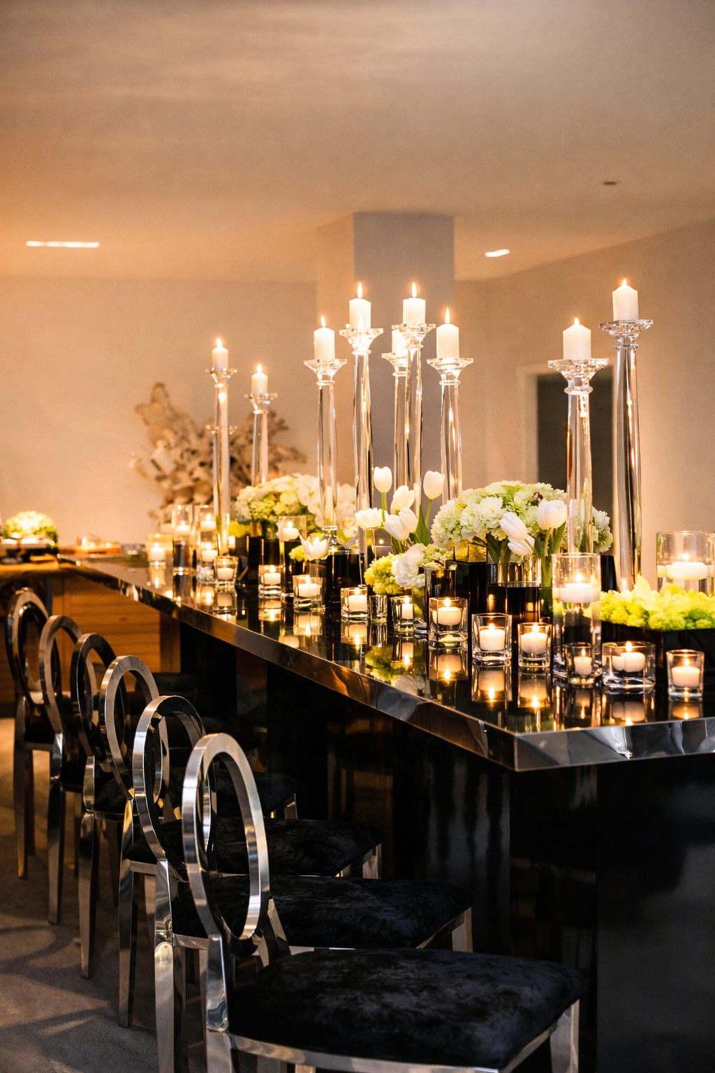 Elegant table setup with tall silver candleholders, white candles, white flowers, and tea light candles on a sleek black table with black chairs.