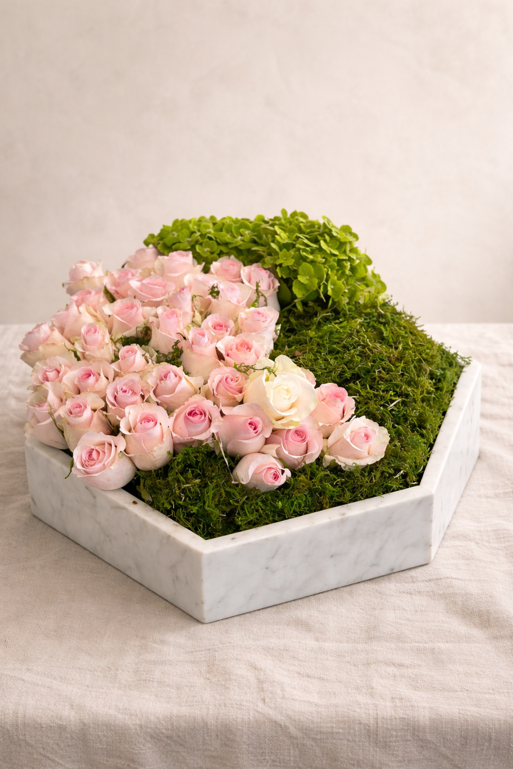 A marble tray with pink and white roses, green foliage, and moss on a beige cloth surface.