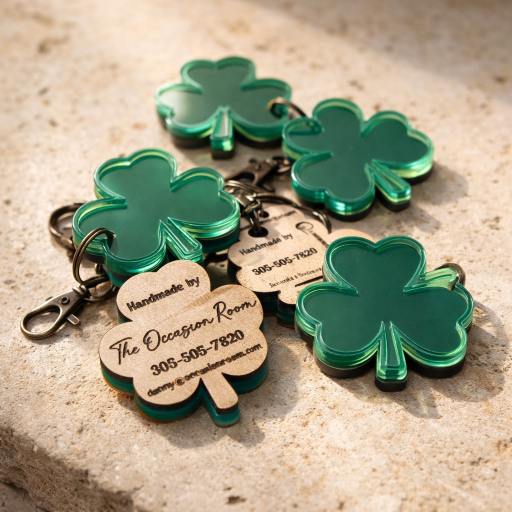 Multiple green shamrock-shaped keychains with tags that read "Handmade by The Oocorgan Room" and contact information, placed on a stone surface.