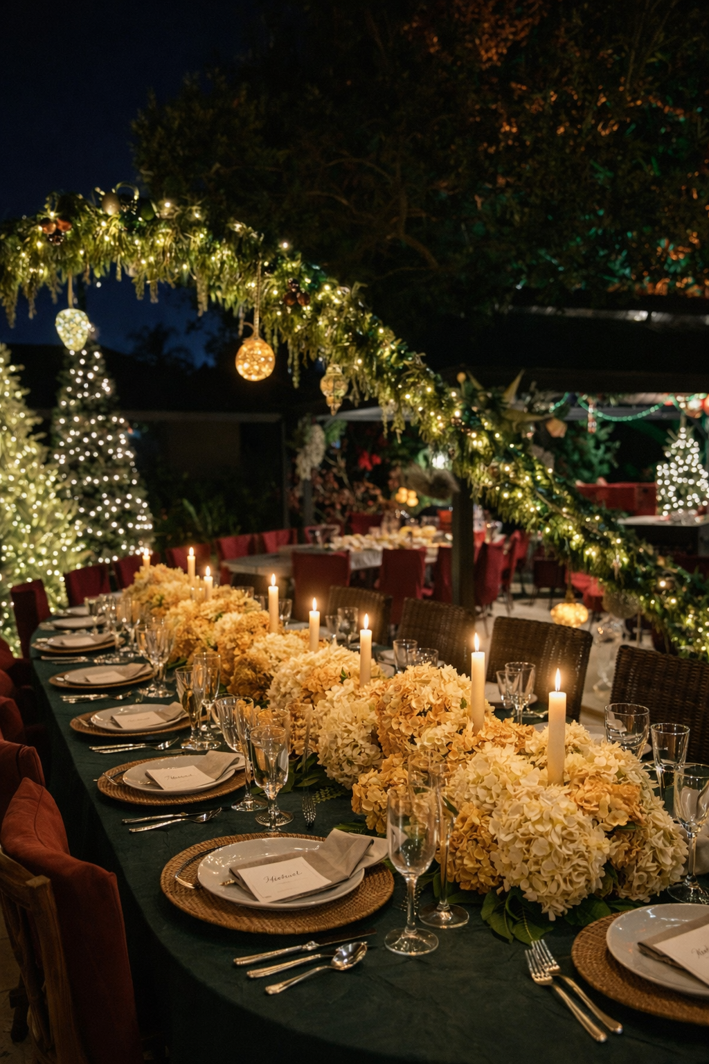 A beautifully decorated outdoor Christmas dinner table at night with floral arrangements, candles, and string lights, surrounded by Christmas trees and holiday decorations.