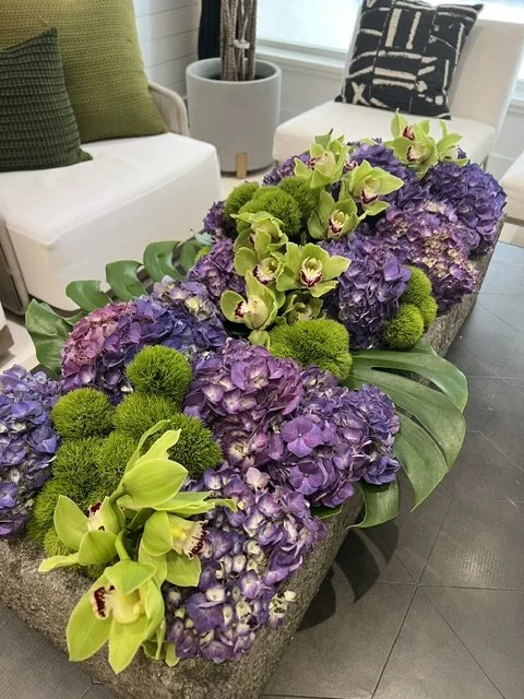 Arrangement of purple hydrangeas, green orchids, and green fuzzy balls in a stone container on a coffee table in a living room with white sofa and black-and-white pillows.
