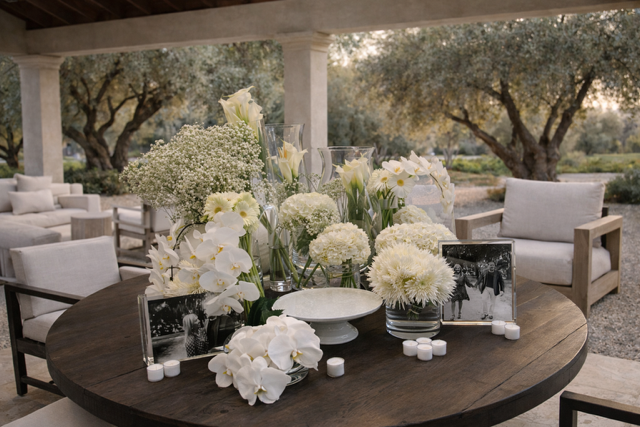 Decorative table setup with white flowers, candles, and framed photos on a dark wooden table, surrounded by outdoor seating and trees.