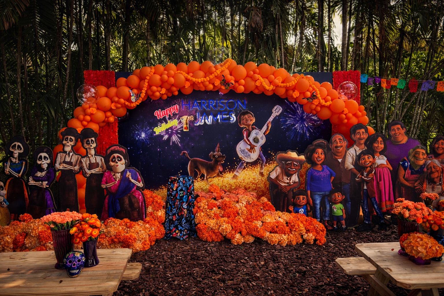 A decorated Dia de los Muertos celebration scene with colorful papel picado, orange marigold flowers, papel cutouts of skeletons, a backdrop with a 'Happy 1st Birthday Harrison James' message, and an animated 'Coco' character playing guitar. The sett
