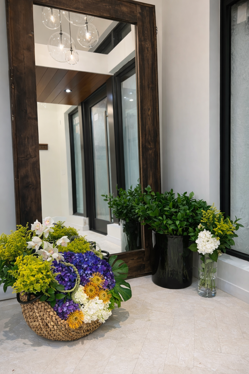 A mirror reflecting a hallway with large windows, a wooden ceiling, and a modern chandelier, with potted green plants and a basket of colorful flowers in the foreground.