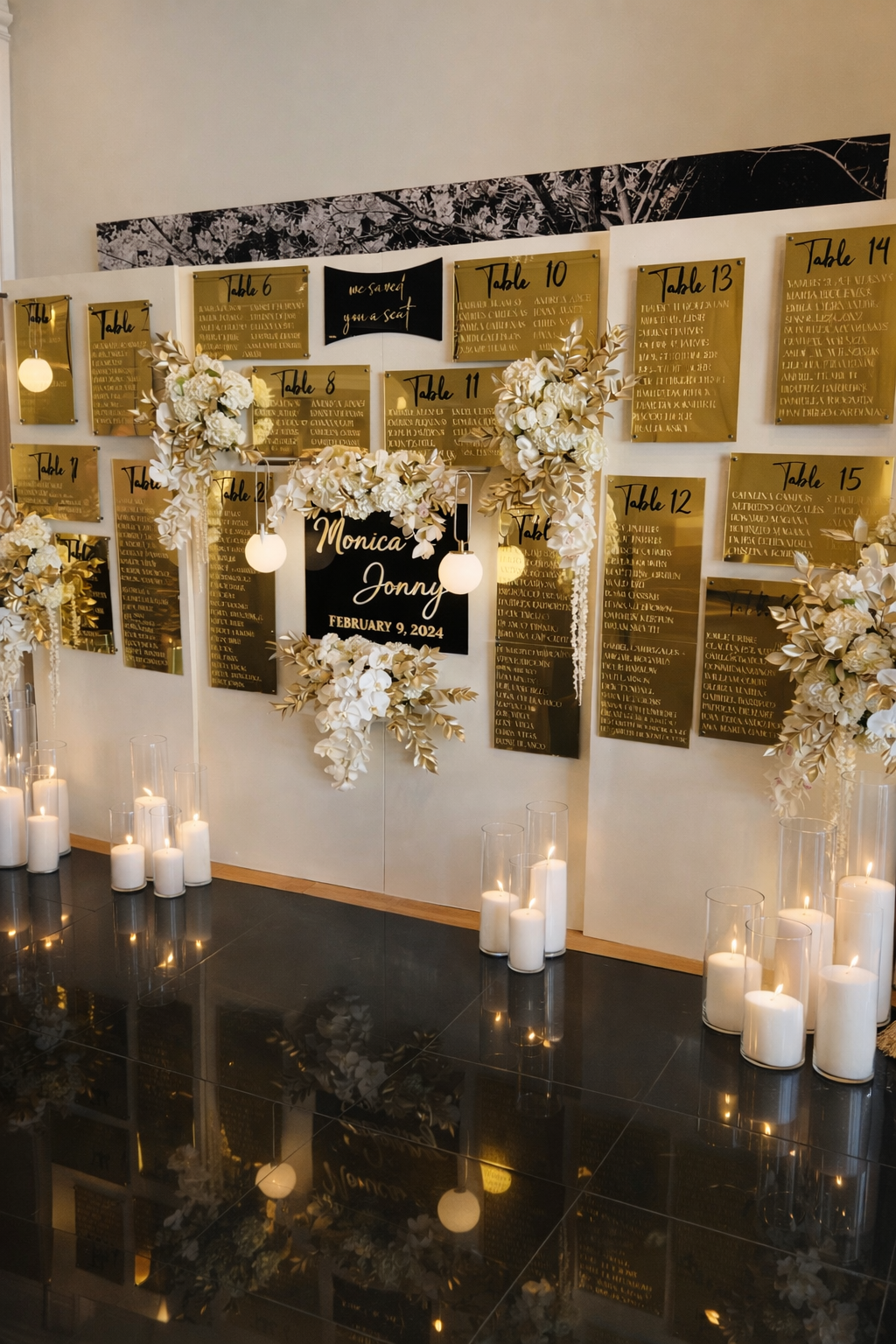 Wedding seating chart with gold and black plaques, floral arrangements, and candles, featuring a black sign with white text that reads 'Monica Jonny February 9, 2024'.