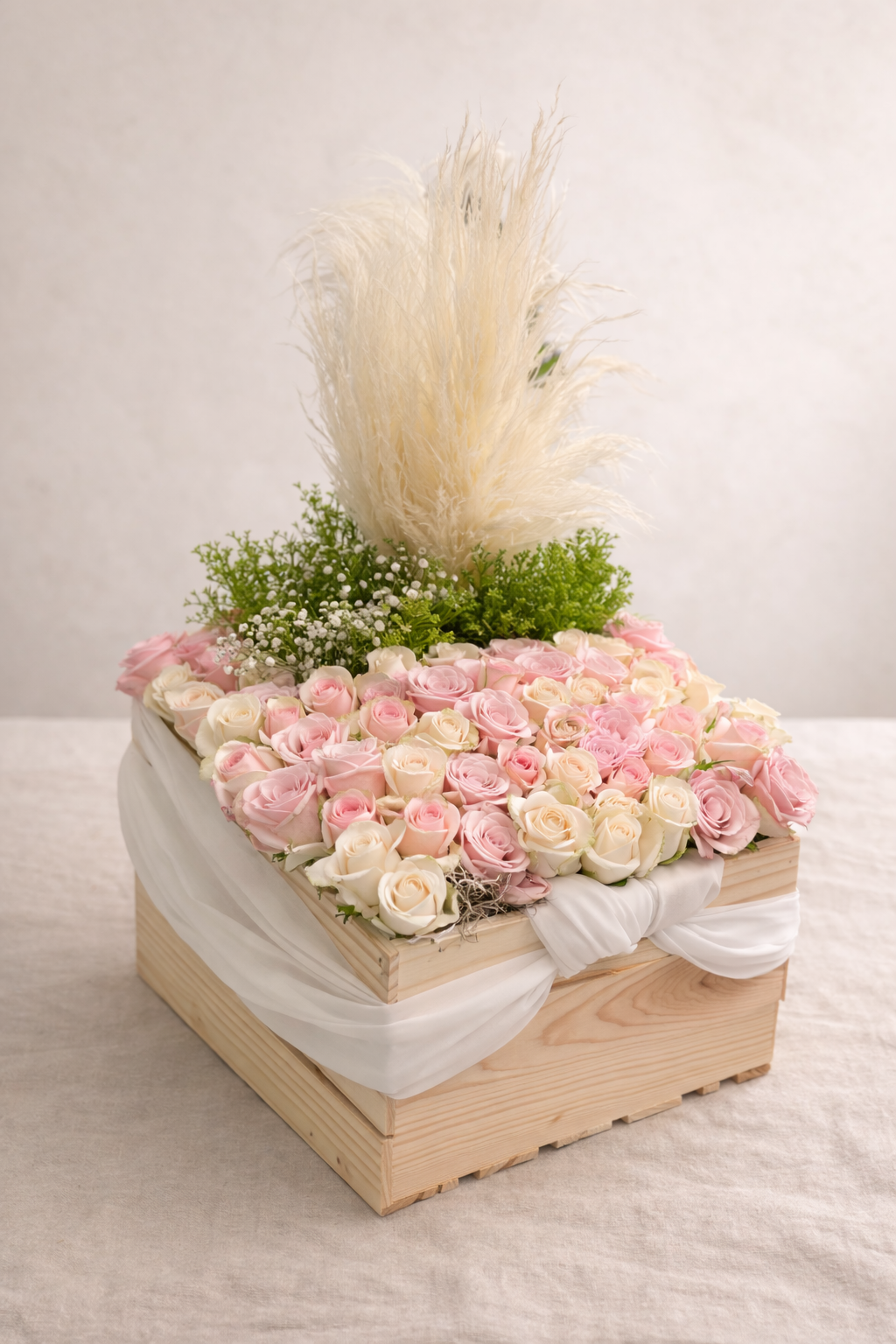 A flower arrangement in a wooden box with pink and white roses, green foliage, and tall beige pampas grass.