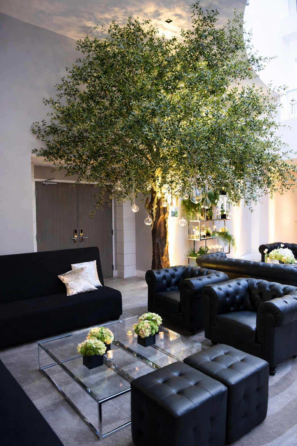 Indoor lounge area with black leather sofas, a glass coffee table with flower arrangements, and a large artificial tree with hanging glass ornaments and warm lighting, set against white walls.