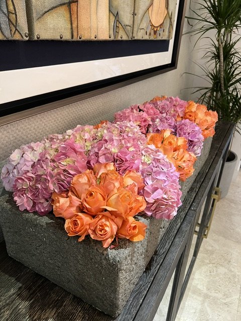 Arrangement of pink, purple, and orange hydrangeas and roses in a gray stone rectangular planter on a dark wooden surface.