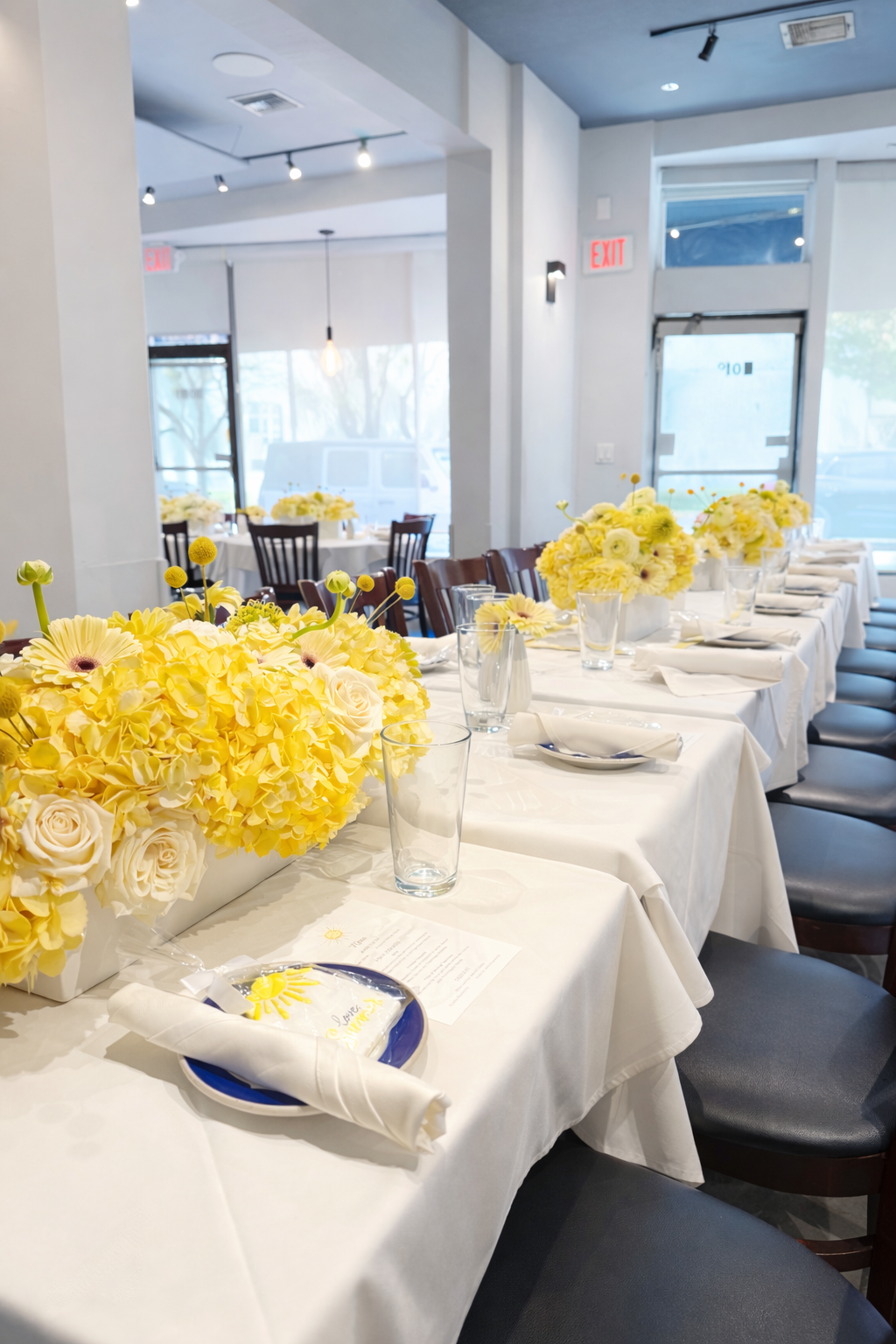 A long dining table set with white tablecloths, yellow floral centerpieces, plates with napkins, and empty glasses in a well-lit restaurant.