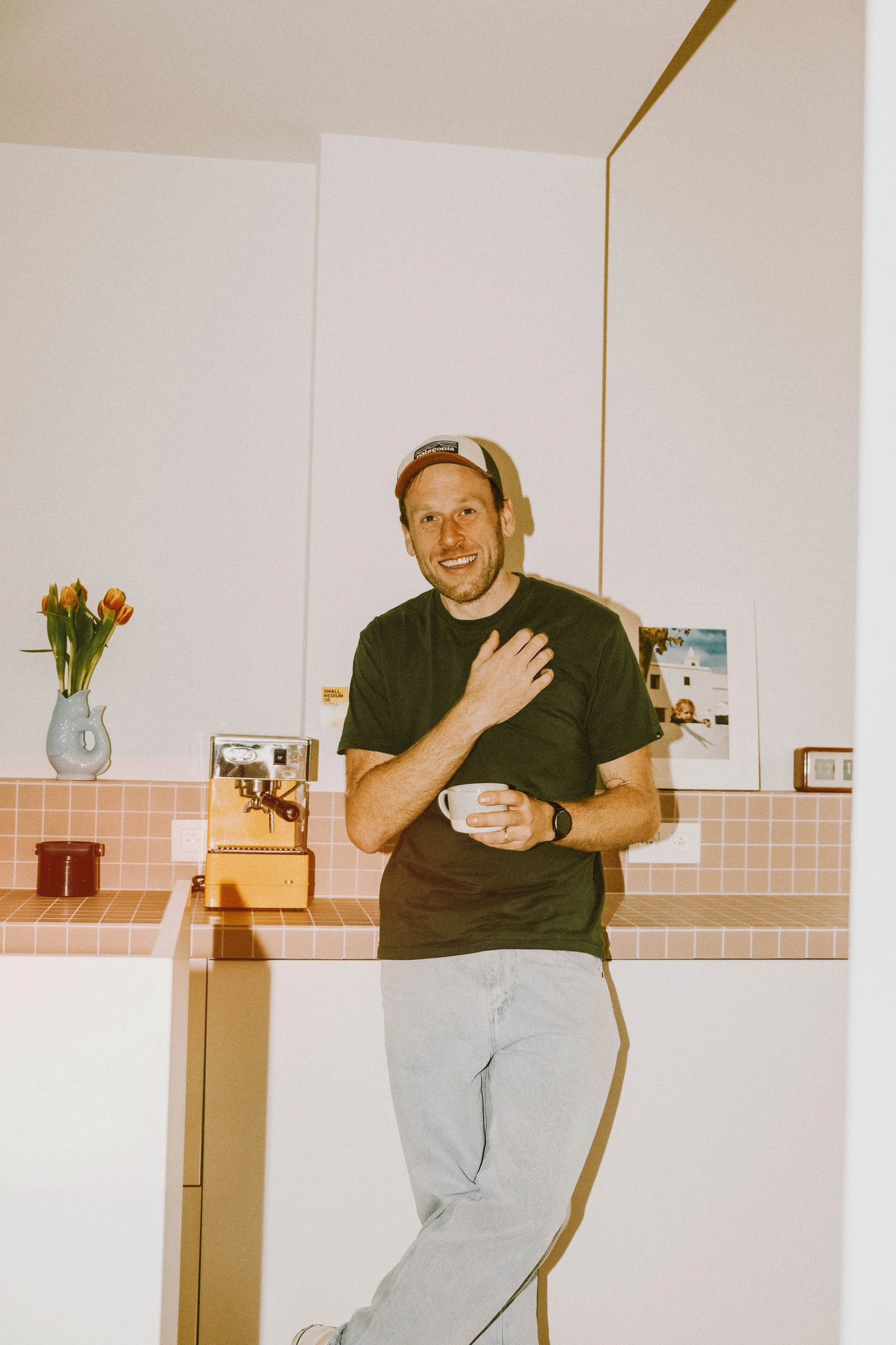 Man in een zwart T-shirt en lichtgrijze broek, lachend met een hand op de borst, houdt een witte mok in de andere hand, in een modern interieur met een vaas met tulpen, een koffiezetapparaat en kunst aan de muur.